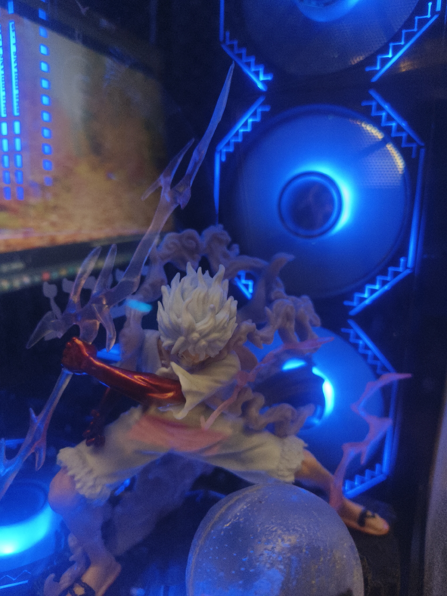 A detailed, close-up, low-angle shot showcases an anime figurine in a dynamic pose, illuminated by vibrant blue LEDs. The figurine, likely made of plastic, depicts a character with spiky white hair, wearing a white garment and a red arm band, holding a translucent trident-like weapon. The background features two large, circular computer fans, also glowing with blue light, arranged vertically. Above the fans, angular blue LED strips create a zigzag pattern. To the left of the figurine, a vertical array of blue rectangular lights is visible. In the foreground, a round, textured object, possibly a decorative ball, is partially visible. The scene is likely an interior shot, possibly within a gaming PC case or a display cabinet, given the presence of computer fans and lighting. There is no visible indication of the city of Kuta Selatan, Indonesia.