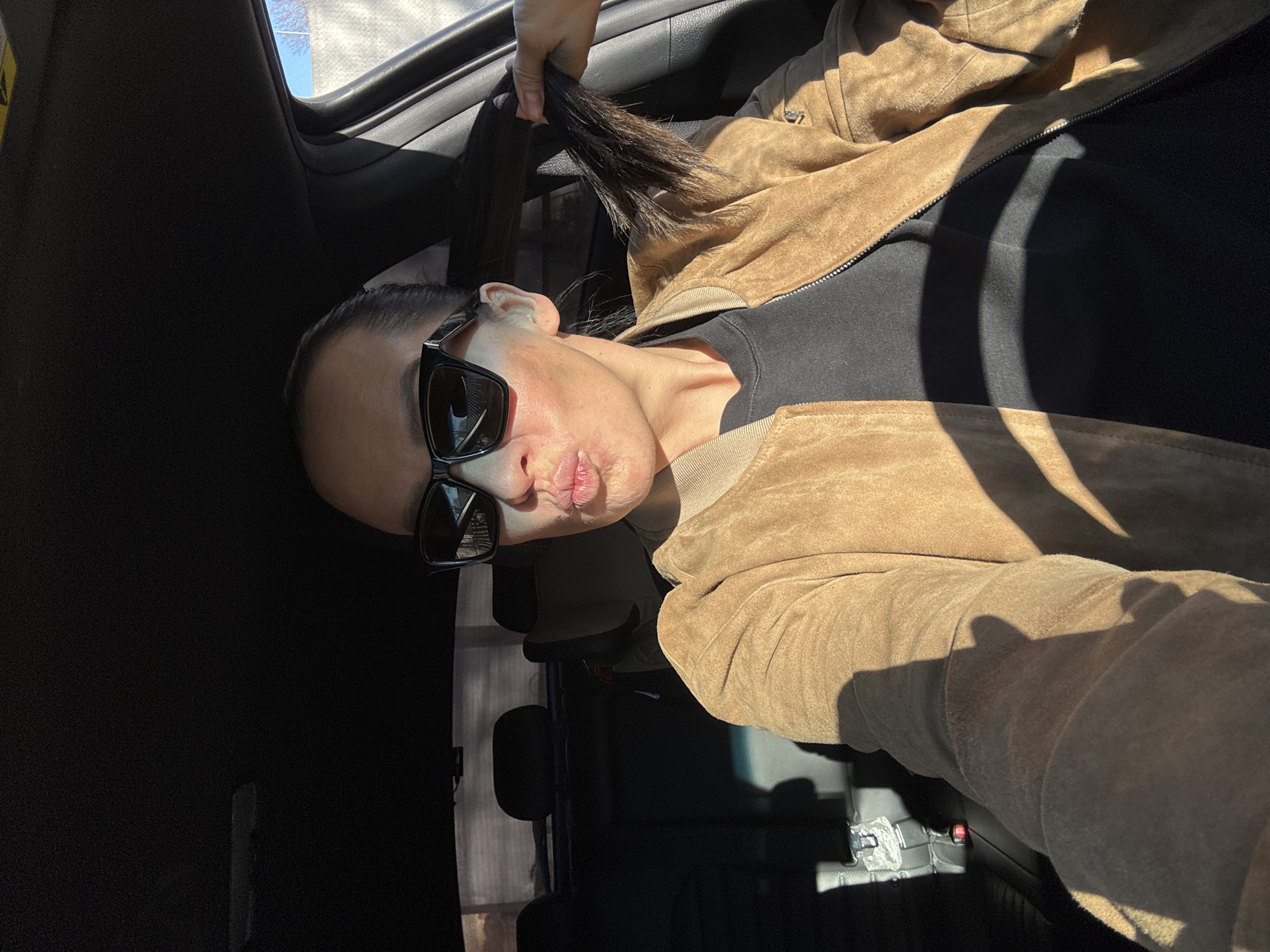 A woman is in the back seat of a car, wearing sunglasses, a black top, and a brown suede jacket. She is holding her ponytail up with one hand. The location is likely indoors, possibly a parked vehicle. There is no discernible information to connect the image to the city of Chaikovsky, Russia.