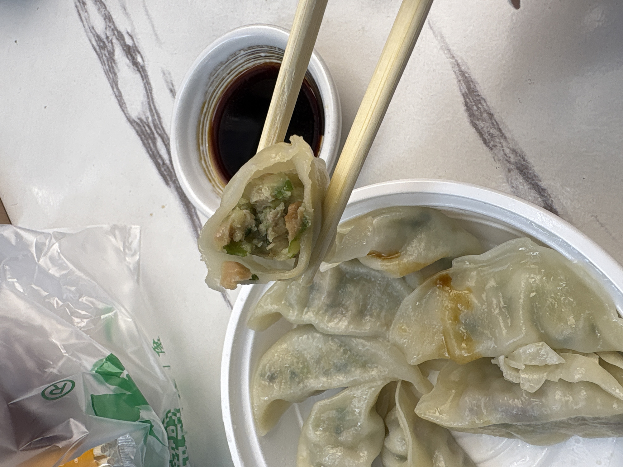 A close-up overhead shot shows a person using chopsticks to pick up a steamed dumpling from a white bowl. The dumpling is filled with green onions and meat. To the right of the bowl is a small dish of soy sauce. The dumplings are on a white marble tabletop with a plastic bag from a restaurant in the background. The logo on the bag indicates it is from Haizhu District, China.