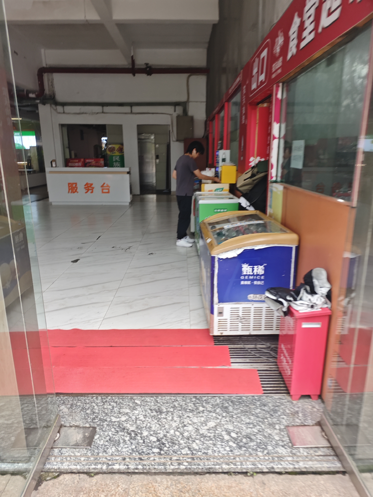 A wide shot captures a casual interior scene possibly in the Chongqing High-tech Zone, China, featuring a service counter and several refrigerators. In the background, a white counter with Chinese text that translates to "Service Counter" is visible, with a person standing behind it. To the right, a storefront with red signage displays Chinese characters, likely indicating a business name. In the foreground, a blue refrigerator with "GEMICE" branding is prominent, stocked with drinks. Adjacent to it, a red trash bin and a stack of clothing are placed on the floor. The environment appears to be a commercial space with tiled flooring.
