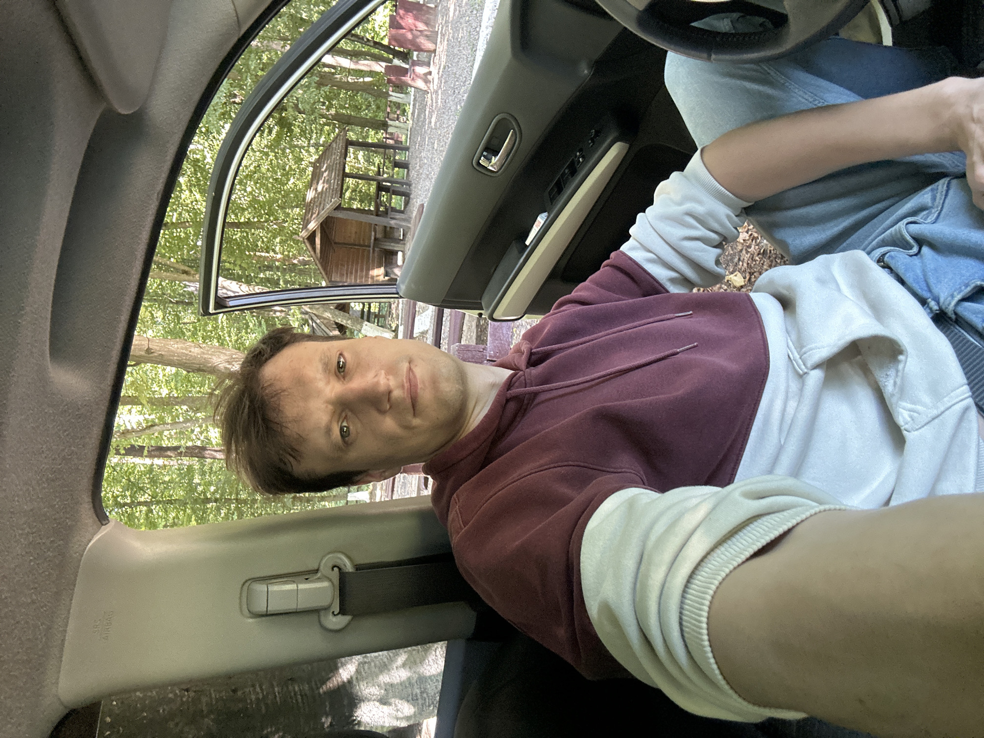 A man is seated in the driver's seat of a car, with the car door open. He is wearing a maroon and white hoodie and blue jeans. The car is parked in a wooded area. In the background, there is a wooden picnic shelter with tables and benches. Trees and foliage are visible throughout the scene. The area appears to be a park or recreational area. The specific location of Shatava, Ukraine is not visually indicated in the image.