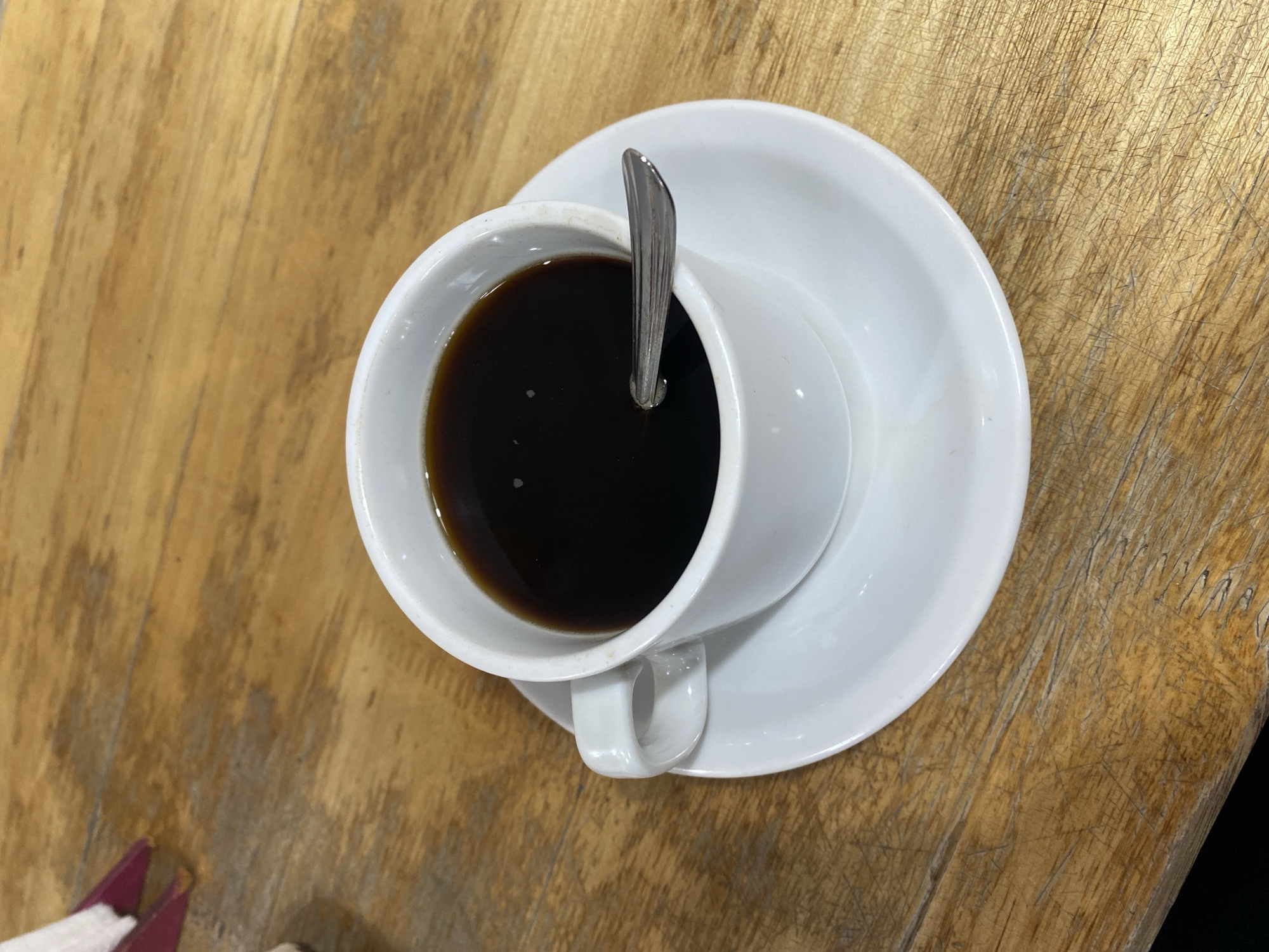 The media file presents an overhead view of a cup of black coffee, resting on a white ceramic saucer. A silver spoon is placed vertically within the dark liquid of the coffee. The cup and saucer are clean and white, providing a stark contrast to the deep, dark brown color of the coffee.
These items are situated on a wooden table, which appears to be light brown or amber in color. The table surface is notably rustic, featuring distinct wood grain patterns and numerous visible scratches and marks, indicating wear and tear. A small, blurred, purple object, possibly a napkin holder or a folded menu, is partially visible in the bottom-left corner of the frame.
The lighting is even, suggesting an indoor setting without harsh shadows. No individuals are depicted in the image. The implied activity is the imminent or ongoing consumption of coffee. No text is visible within the frame. The context for this scene is Monte Grande, Argentina.