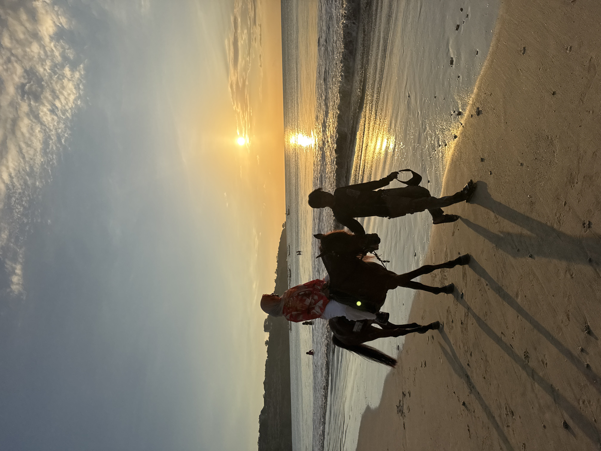 A silhouetted figure walks on the beach next to a horse being ridden by a person wearing a white top and head covering, and a red and white patterned shirt. The setting is at sunset, with the sun reflecting off the ocean and wet sand. The location appears to be in Kuta Selatan, Indonesia.