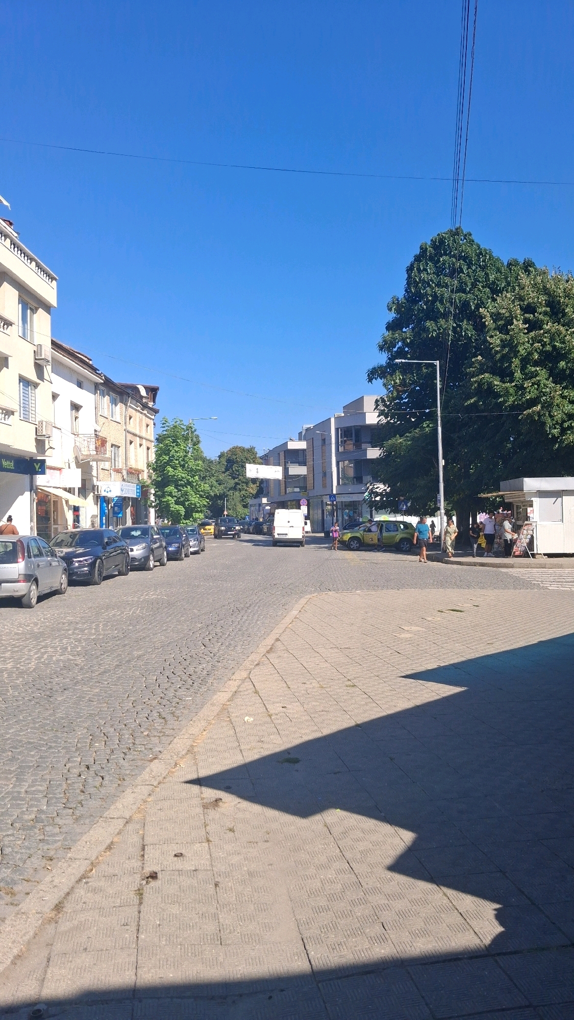 The image depicts a street scene in the city of Gotse Delchev, Bulgaria, under a clear blue sky on a sunny day. The street is paved with cobblestones on the left side, leading into a sidewalk with square tiles on the right. Several cars are parked along the cobblestone street. Buildings line the left side of the street, featuring architectural styles that suggest a mix of older and newer constructions. On the right side, modern buildings and a small kiosk are visible, with trees providing shade. Several people are present, walking on the sidewalk or near the kiosk. The overall impression is of a typical urban environment in a European city.