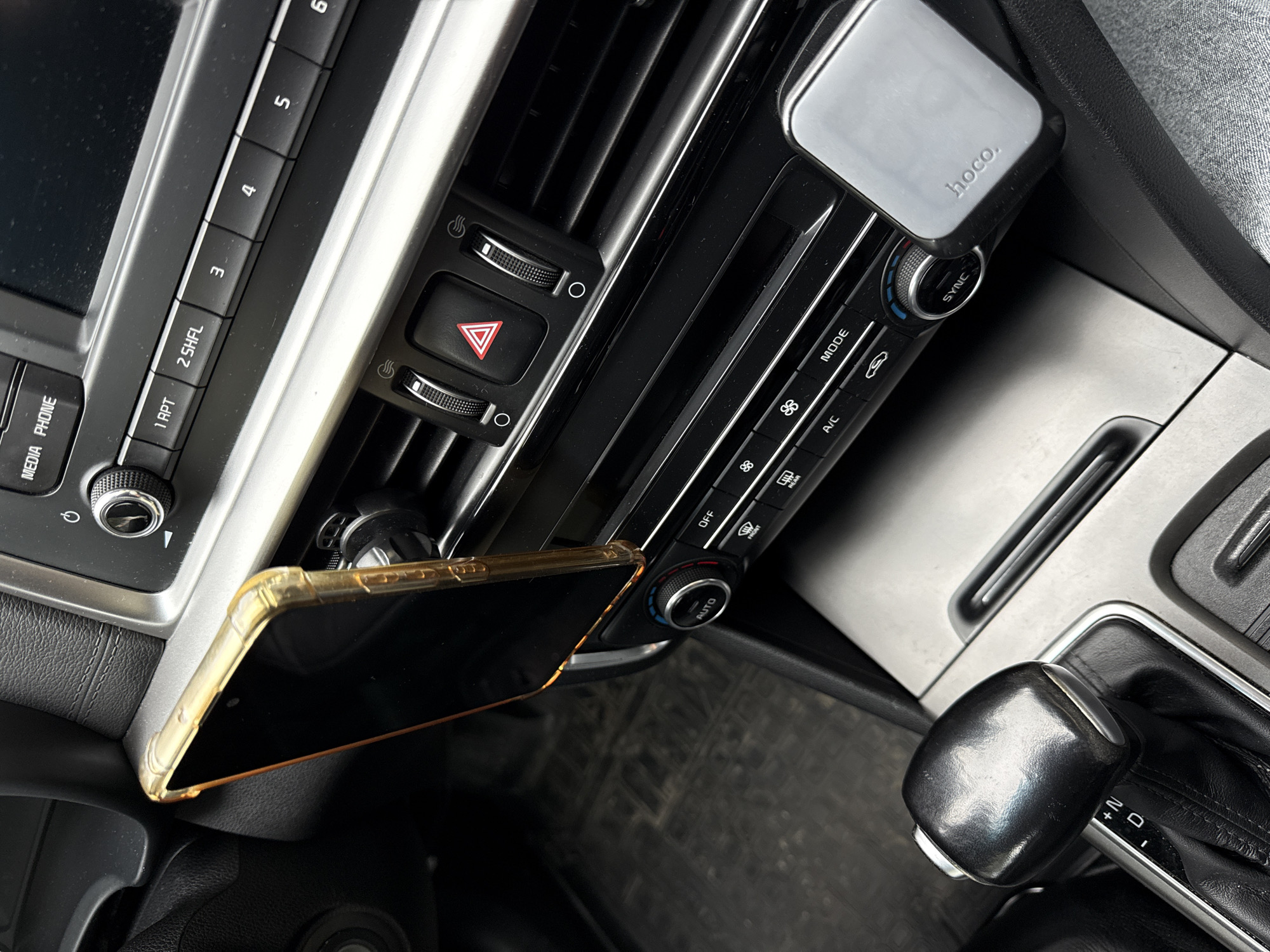 The image shows the interior of a car, specifically the center console area. A smartphone in a yellow case is mounted on the left air vent. Next to it is the car's audio system controls and hazard light button. To the right of the center console is the automatic transmission gear shift. On the upper right is an air vent phone mount with the brand name "hoco" printed on it. Below it are the car’s climate controls.
There's no visible evidence in the image to indicate a specific location in Sochi, Russia or the presence of people or specific actions or interactions.
