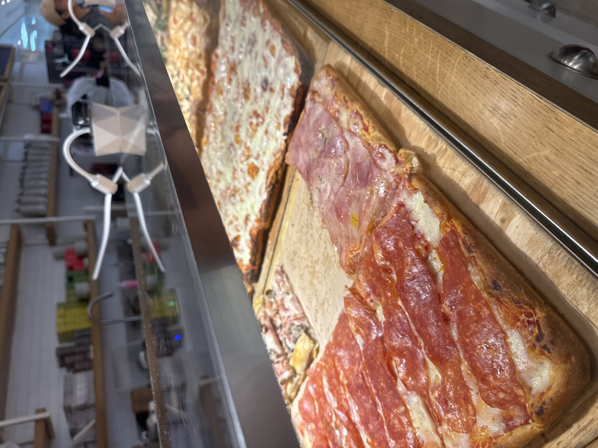 The image shows two types of rectangular pizzas displayed in a glass-enclosed case. The first pizza features a layer of melted cheese. The second pizza has sections topped with different meats, including salami and ham. Both pizzas are placed on wooden boards, which sit inside the display case. The case itself has a metallic frame and appears to be located in a deli or restaurant setting. The image's location is Aventura, United States of America.