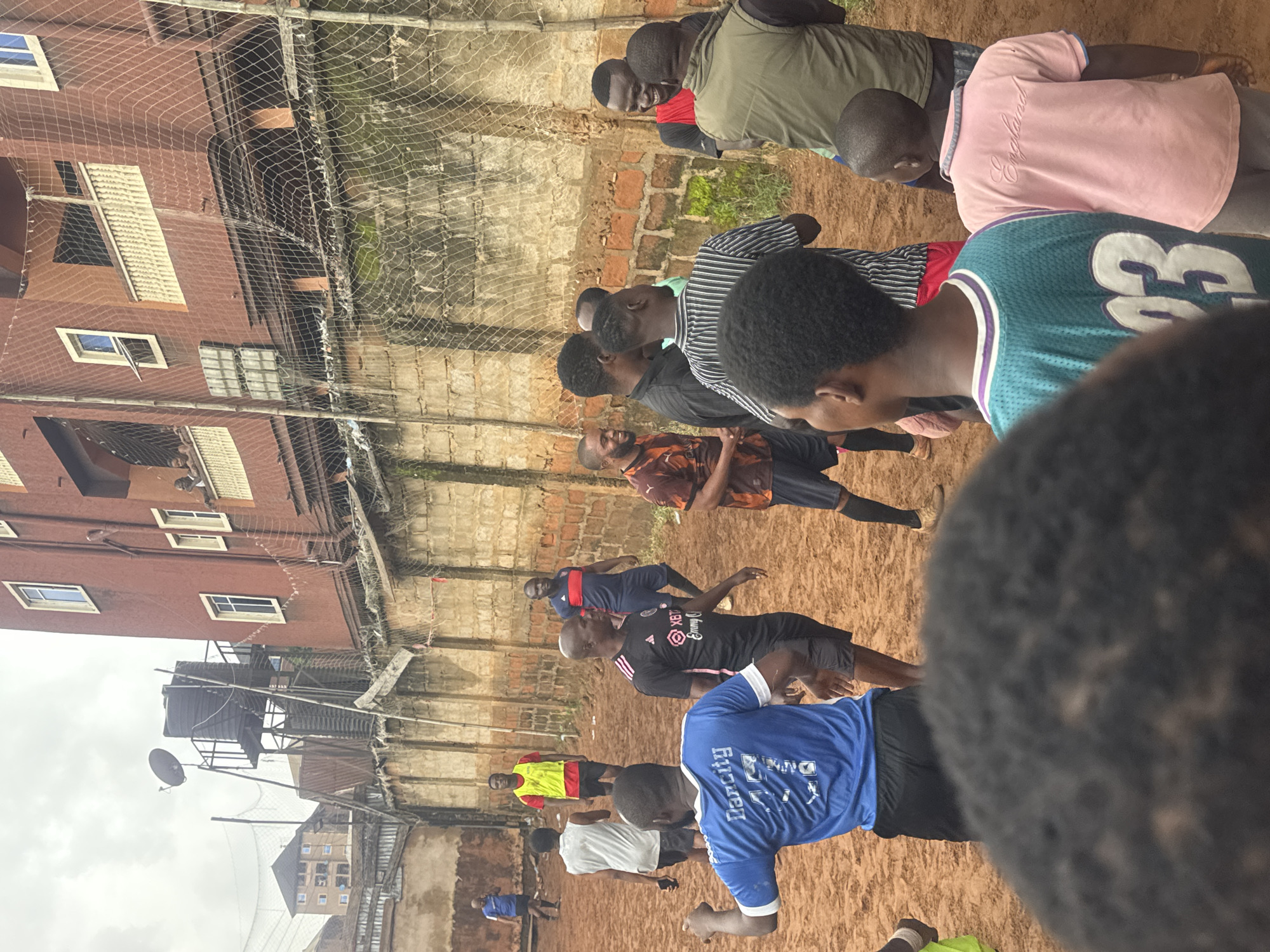 In Nkpor, Nigeria, a group of men are gathered on a dirt football field, playing a match under an overcast sky. The field is surrounded by a brick wall topped with a net, and several multi-story buildings are visible in the background, including a reddish-brown residential building with balconies and windows. Some players are in athletic wear, including jerseys and shorts, while others wear casual clothing. One man in a blue jersey with the name "Dancity" and the number "11" is visible from behind, facing away from the camera. Another player in a black and pink striped shirt is seen from the side. A young boy in a pink shirt with "England" written on it stands near the front of the group. The overall scene depicts a casual football game taking place in an urban environment.