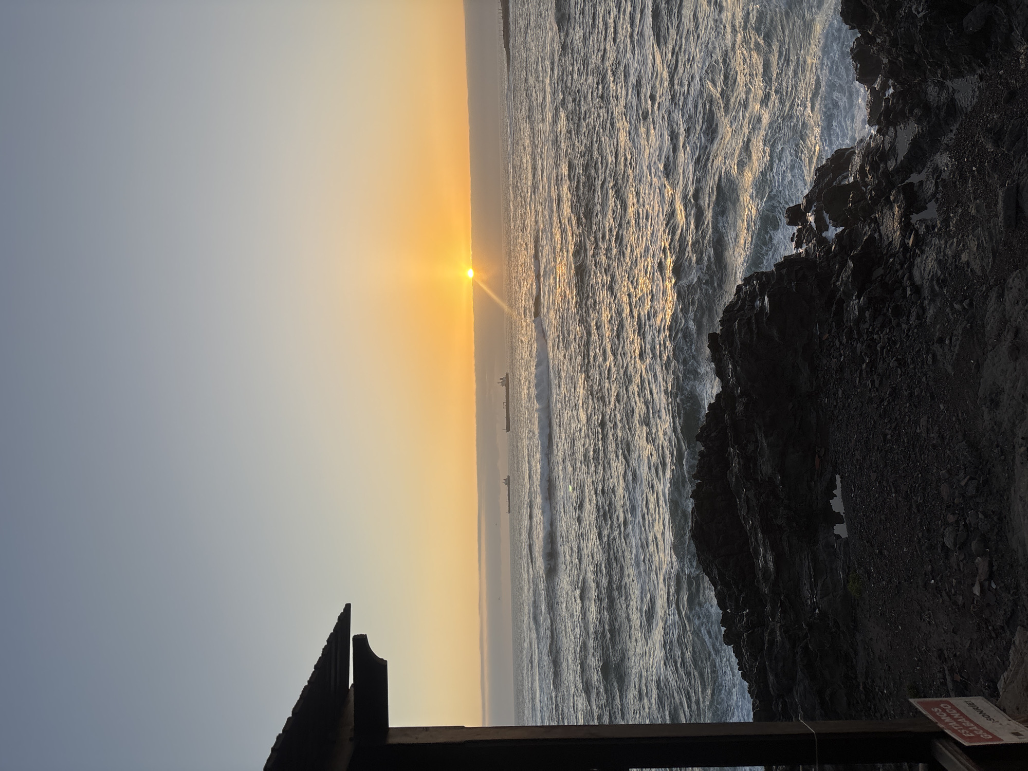 The image depicts a sunset over the ocean in Arica, Chile. Two ships are visible on the horizon, silhouetted by the setting sun. The ocean is choppy, with waves crashing against a rocky shoreline on the right. In the foreground, a dark, angular structure suggests a man-made barrier or observation point. The sky transitions from a light blue to a warm orange glow near the horizon.