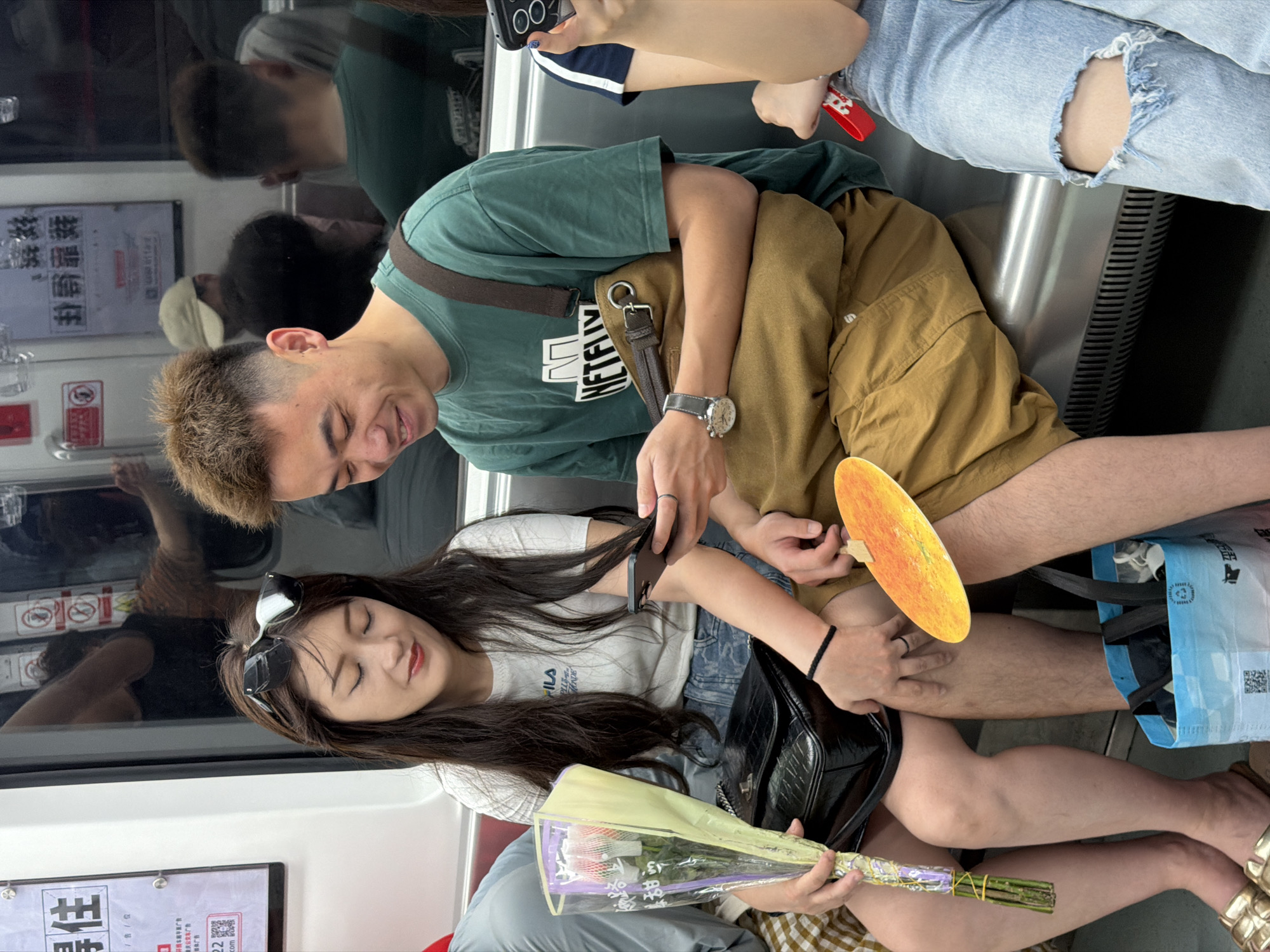 On a subway car, a man and a woman are seated next to each other. The man, wearing a green t-shirt with "NETFLIX" printed on it and brown shorts, is looking down at his phone. He has short, spiky brown hair and is wearing a watch on his left wrist. The woman, wearing a white t-shirt and denim shorts, is also looking at her phone. She has long dark hair with sunglasses perched on top of her head. In her lap, she holds a bouquet of pink and white flowers wrapped in clear plastic and a black handbag. A circular orange fan lies on the seat between them, and a blue reusable shopping bag is on the floor. The subway car interior features a metallic bench seat, posters on the wall, and reflections in the window. The scene appears to be in Yuzhong District, China.