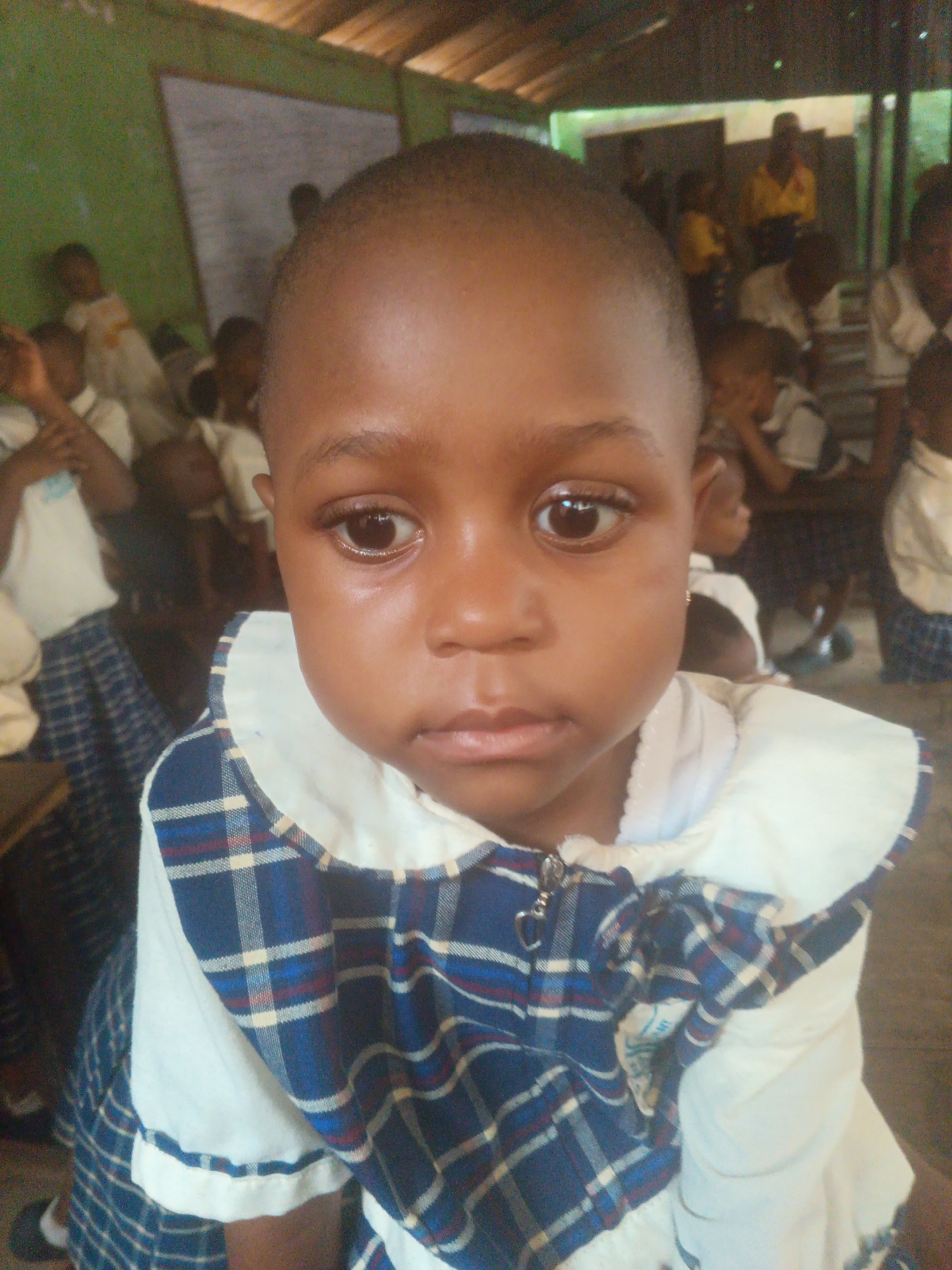 A close-up, slightly grainy photograph shows a young Black child, likely a girl, in the foreground. She has dark skin, short hair, and is looking directly at the camera with a somber expression, her eyes slightly watery. She is wearing a white shirt with a blue and red plaid collar and accents. The background is blurred and dimly lit, showing other children and adults in what appears to be a classroom or gathering space. The ceiling is visible, constructed of wooden planks. The overall impression is that of a candid moment captured in a community setting, potentially a school or church in Onitsha, Nigeria.