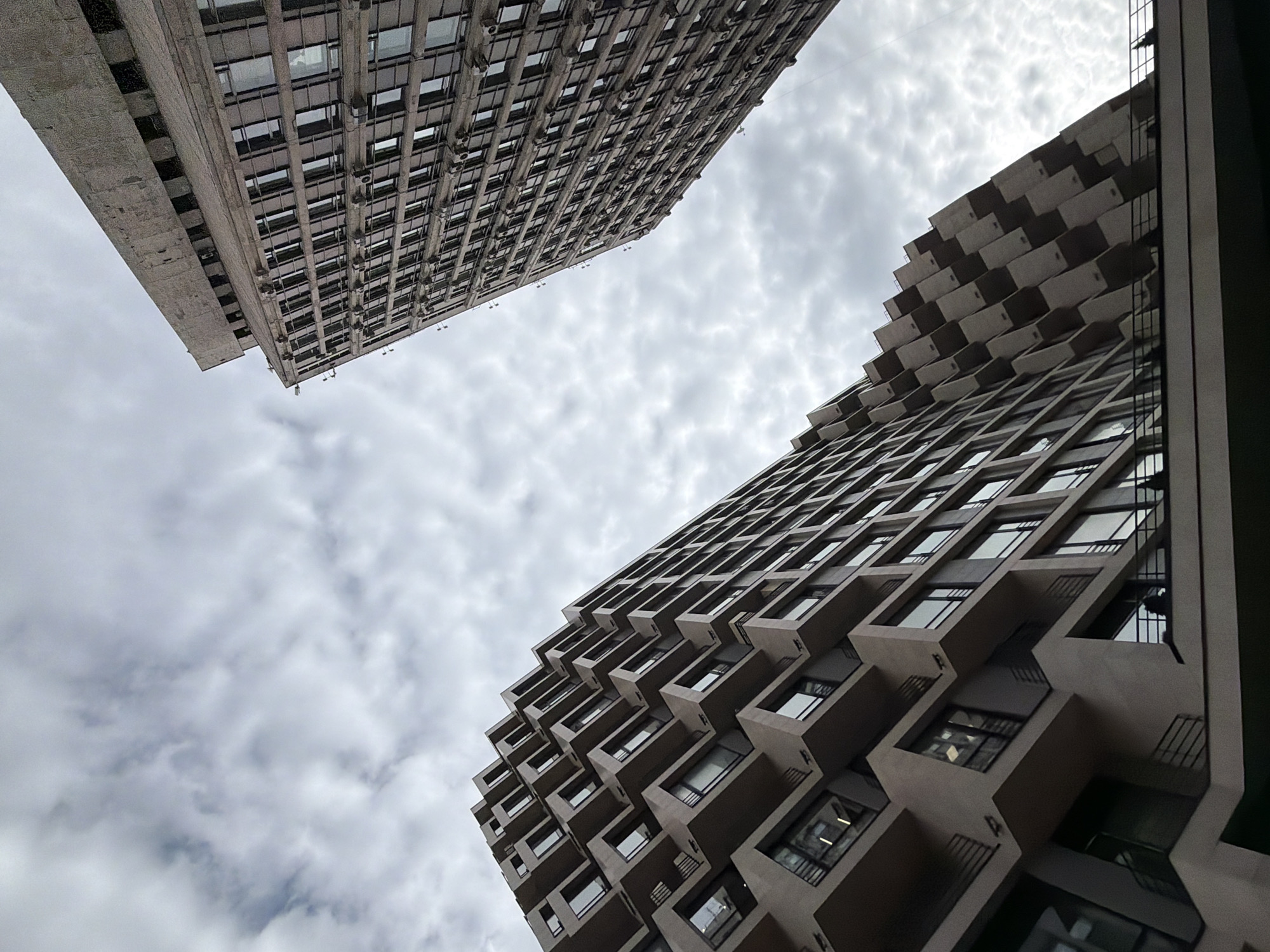The image displays two Brutalist-style buildings viewed from a low angle, looking upwards towards a cloudy sky. The building on the left is a long, rectangular structure with a facade composed of numerous windows and visible external air conditioning units. Its concrete exterior appears weathered, with some discoloration. The building on the right has a more distinctive design, featuring a staggered, layered facade with recessed windows that create a patterned effect. Both buildings are tall and imposing.
The sky is overcast with scattered clouds, suggesting it is daytime, likely a cool or temperate day. There are no people clearly visible in the frame, and no specific activity is depicted; the scene focuses solely on the architecture and the sky. The stark, geometric forms of the buildings, combined with the muted lighting due to the clouds, evoke a sense of urban grandeur and perhaps a slightly melancholic or somber atmosphere. The architectural style and the implied urban environment are consistent with a location like Moscow, Russia, known for its substantial Soviet-era architecture. No visible text is present in the image.