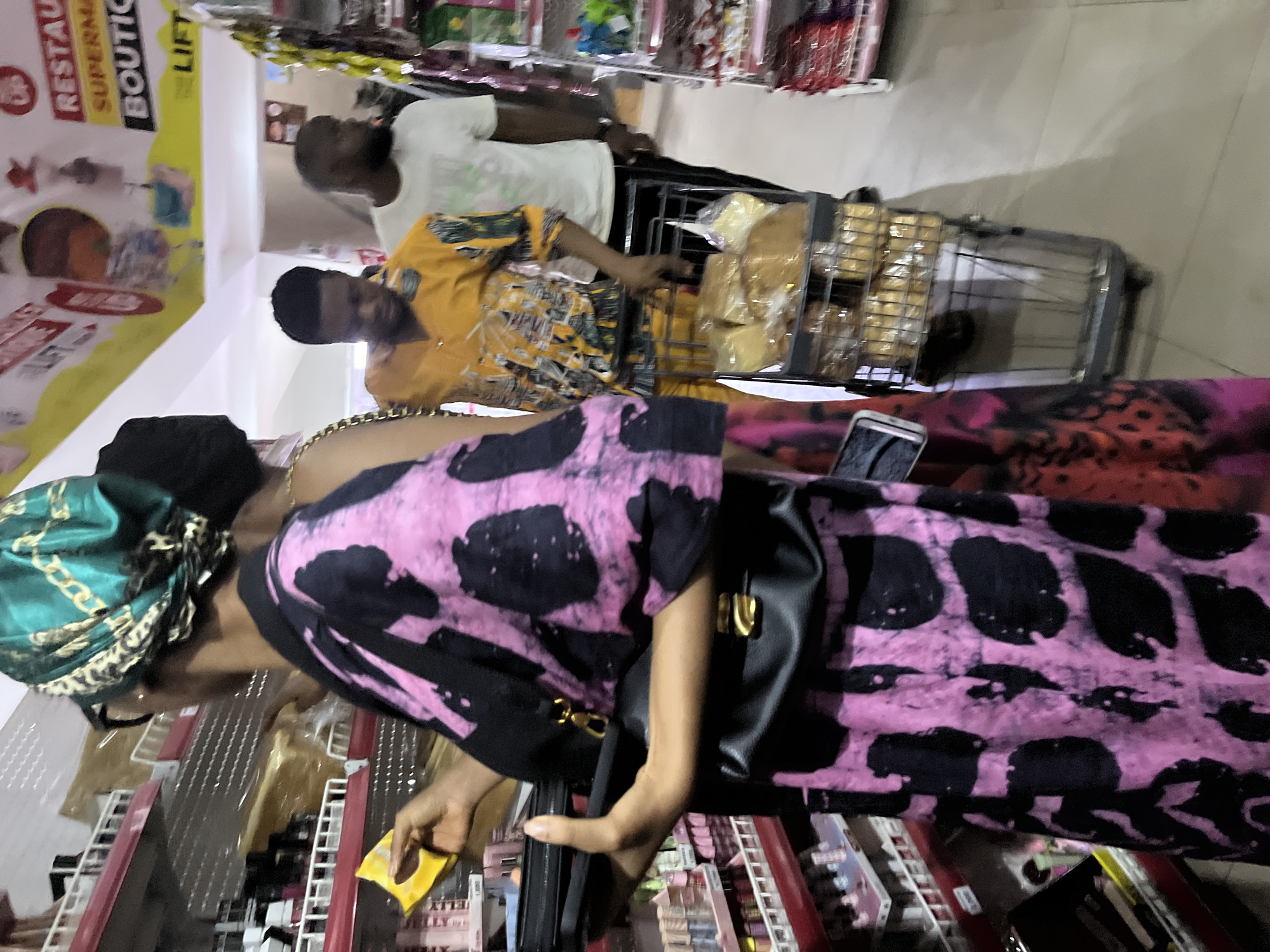 A woman in a pink and black patterned outfit and a green headwrap is standing in a supermarket aisle in Abuja, Nigeria, examining products on a shelf. She is holding a yellow item and has a black purse on her shoulder. In the background, two men are pushing a shopping cart filled with loaves of bread. A supermarket sign is visible in the upper left corner of the image.