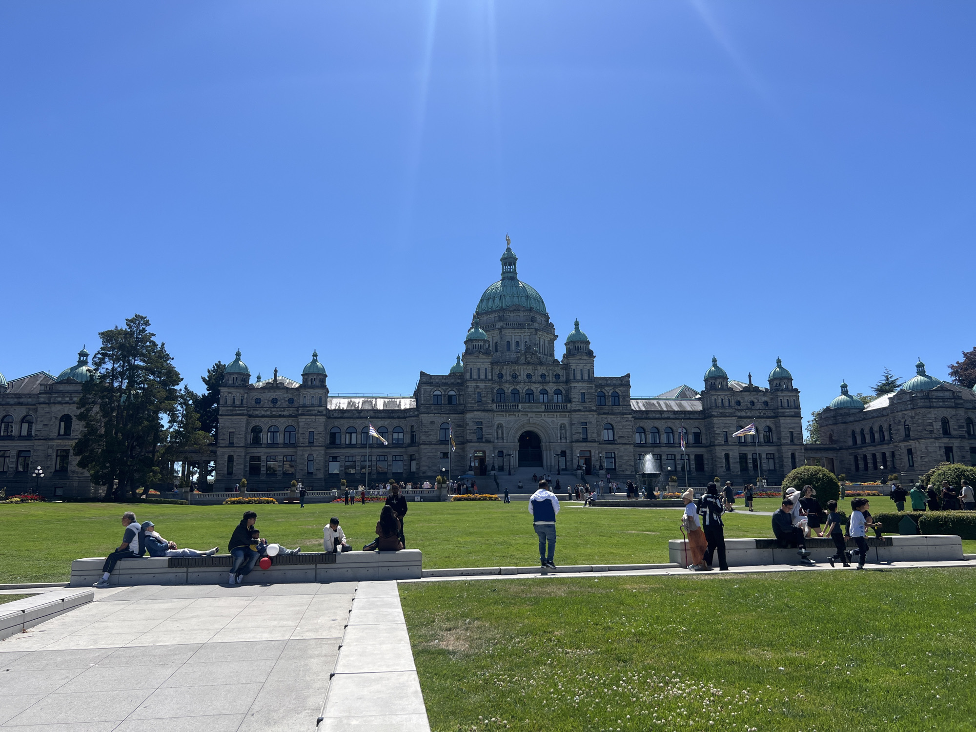 A wide shot captures the majestic facade of the British Columbia Parliament Buildings in Victoria, Canada, on a bright, sunny day. The imposing stone structure, adorned with green domes and intricate architectural details, dominates the background under a clear blue sky. Sunlight glints off the building and the sky, indicating mid-day.
In the foreground and middle ground, a lush green lawn is dotted with small white clover flowers. Several groups of people are scattered across the lawn and sitting on low, light-colored stone benches. On the far left, a group of three people are casually seated, with one person lying down, suggesting a relaxed atmosphere. Closer to the center, a family sits together, with a child held by an adult, and a red and white balloon visible. Other individuals and small groups are also seen enjoying the outdoors, some standing, others seated.
Towards the center of the image, a large fountain sprays water, adding to the lively scene. Flags fly from poles in front of the Parliament Buildings. The overall mood is one of leisure and enjoyment of a beautiful day in a public space. The architecture, the setting, and the presence of visitors suggest this is a popular tourist destination. No visible text is present in the image.