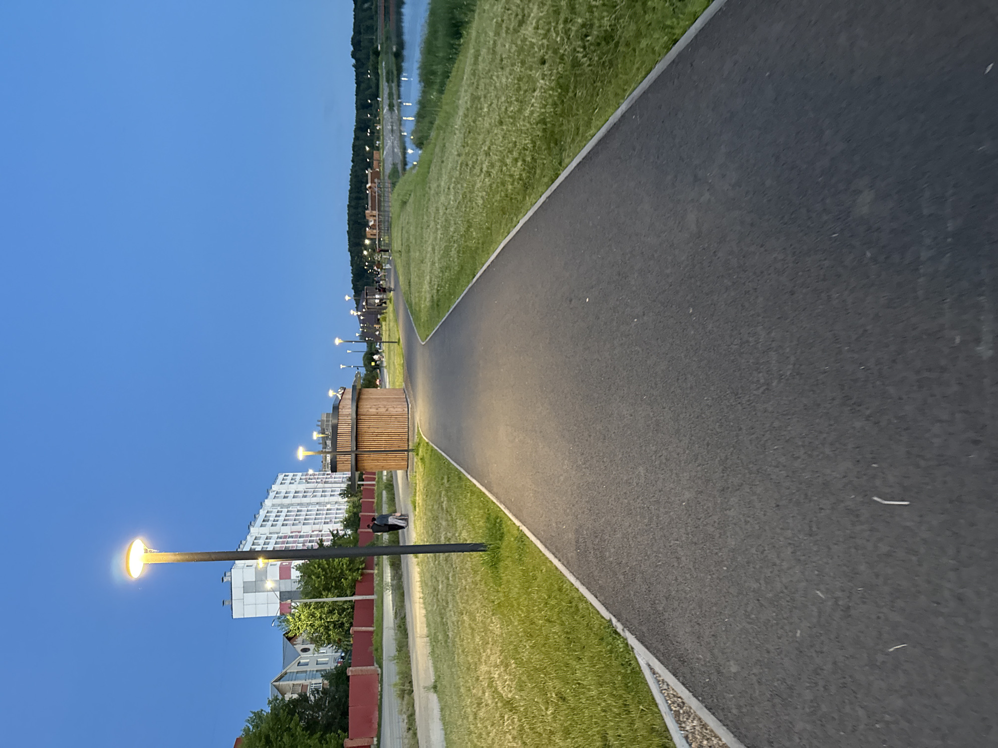A paved pathway curves through a grassy park area next to a body of water. The sky is a deep, clear blue, indicating either early morning or evening. Several lampposts illuminate the pathway, casting a warm glow on the asphalt. In the distance, trees line the shore of the water, and beyond them, a few buildings are visible. Closer to the pathway, there's a modern wooden structure, possibly a rest area or pavilion, and a few benches. A tall, white apartment building with red and white trim stands to the left of the pathway, surrounded by trees and smaller houses. The scene appears peaceful and serene, suggesting a quiet evening stroll or a moment of relaxation in the park. There is no visible text in the image.