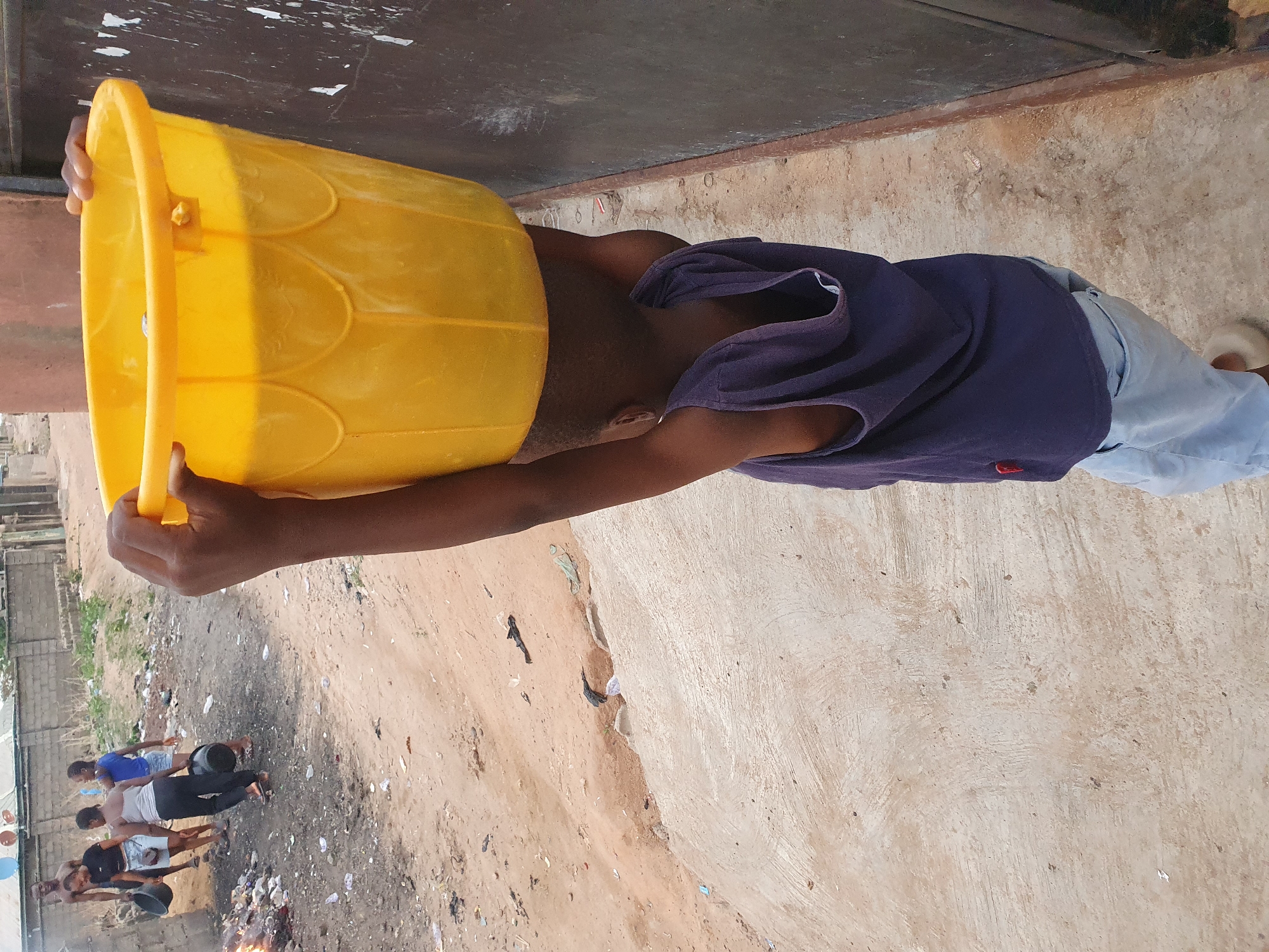 The image, rotated 90 degrees clockwise, captures an outdoor scene in Lokoja, Nigeria, under bright daylight. The central figure is a dark-skinned person, likely a young man or boy, holding a vibrant yellow plastic bucket over their head, completely obscuring their face and upper chest. They are wearing a dark blue garment, possibly a t-shirt or a towel draped over their shoulders, and light blue or grey trousers, with a light-colored sandal visible on their right foot. Their hands are gripping the bucket's handle from underneath.
The ground in the foreground transitions from rough, light brown dirt or sand with scattered debris to a smoother concrete surface. In the mid-ground and background, several other individuals, appearing to be young adults or teenagers, are gathered. One person is visible holding a dark bucket, while another stands near a small, smoldering fire producing light smoke, suggesting communal activity, possibly involving waste burning or cooking. Structures like a grey block wall and green fencing are visible in the far left background. The bright, direct sunlight indicates it is daytime with clear weather. No discernible text is visible.