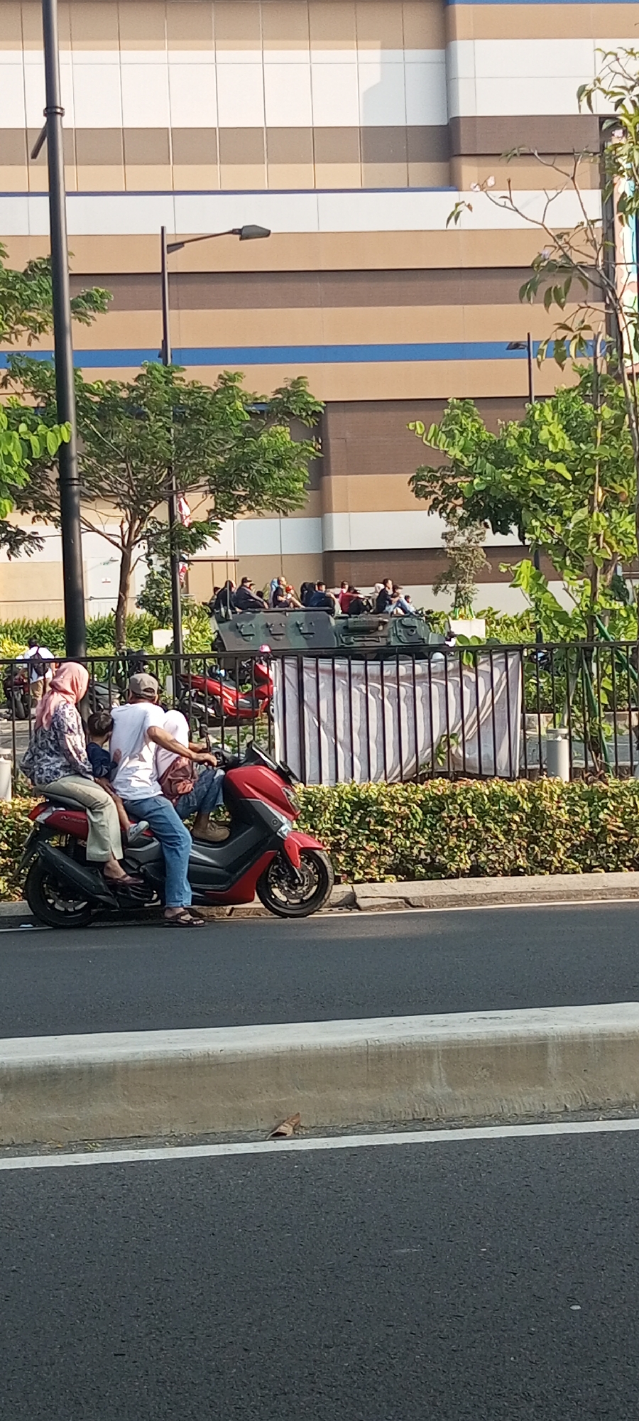 In Lambangjaya, Indonesia, on a sunny day, a red scooter is parked on the side of a road with a concrete barrier. The scooter has three occupants: a woman wearing a pink hijab and a patterned blouse, a man in a white t-shirt and blue jeans riding the scooter, and a child seated between them. In the background, a military vehicle with people sitting on top is visible behind a black metal fence. The vehicle appears to be camouflaged. A building with horizontal stripes of white, brown, and blue is situated behind the military vehicle, and there are trees and some shrubbery in the foreground and middle ground. A lamppost is also present to the left of the frame.