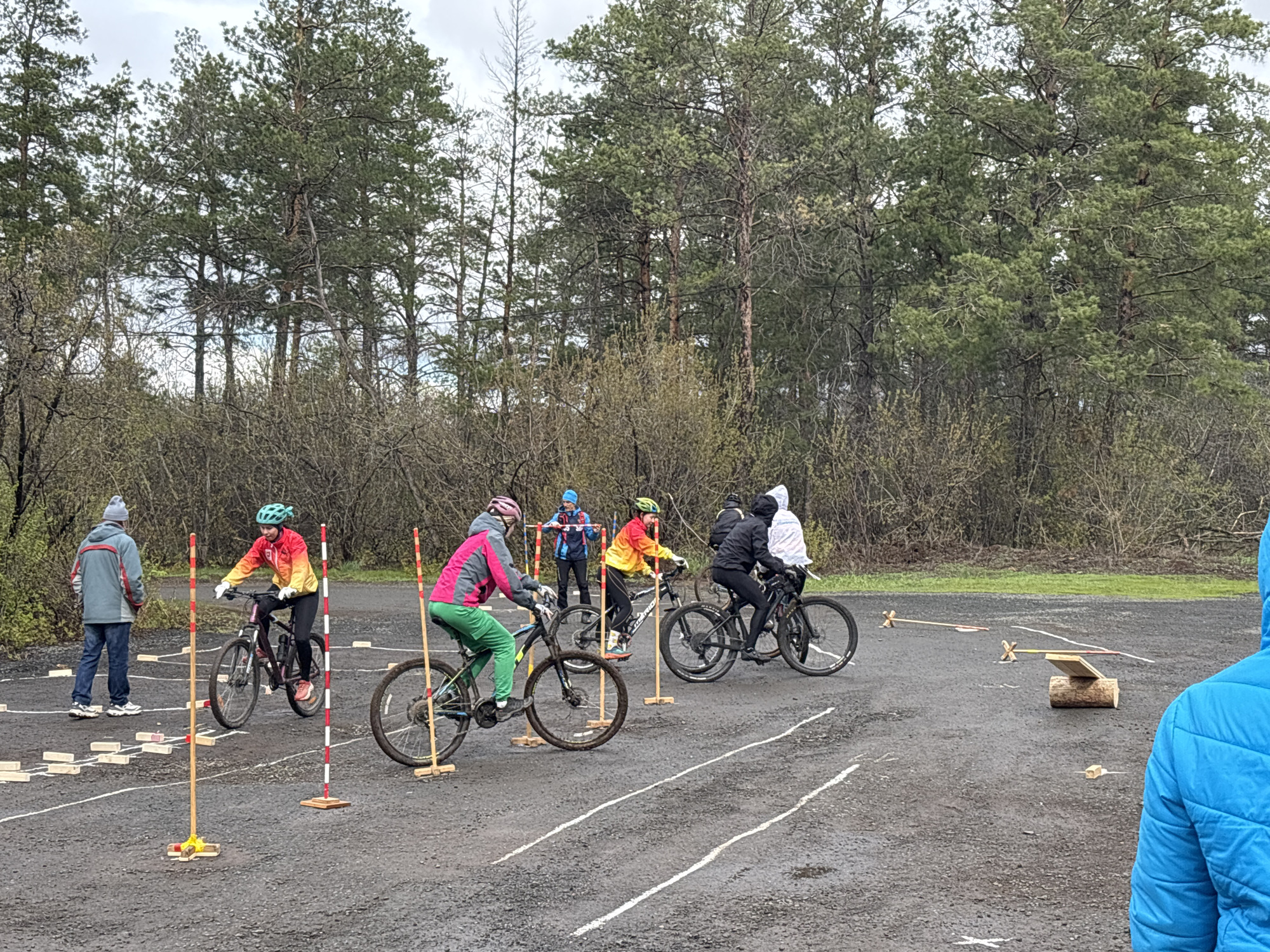 In an outdoor asphalt area, possibly a parking lot or training course, several people are riding bicycles through a marked obstacle course. The course is defined by painted white lines, wooden blocks, and vertical poles on wooden bases. Five cyclists are visible, each wearing helmets and casual outdoor clothing, maneuvering their bikes through the course. There are also at least two other people, one appearing to be an observer standing near one end of the course and another partially visible wearing a blue jacket. The course also includes a raised wooden plank balanced on a log. The backdrop consists of a dense collection of trees, indicating a park or wooded area. There is no discernible information to indicate it is the city of Orenburg, Russia.