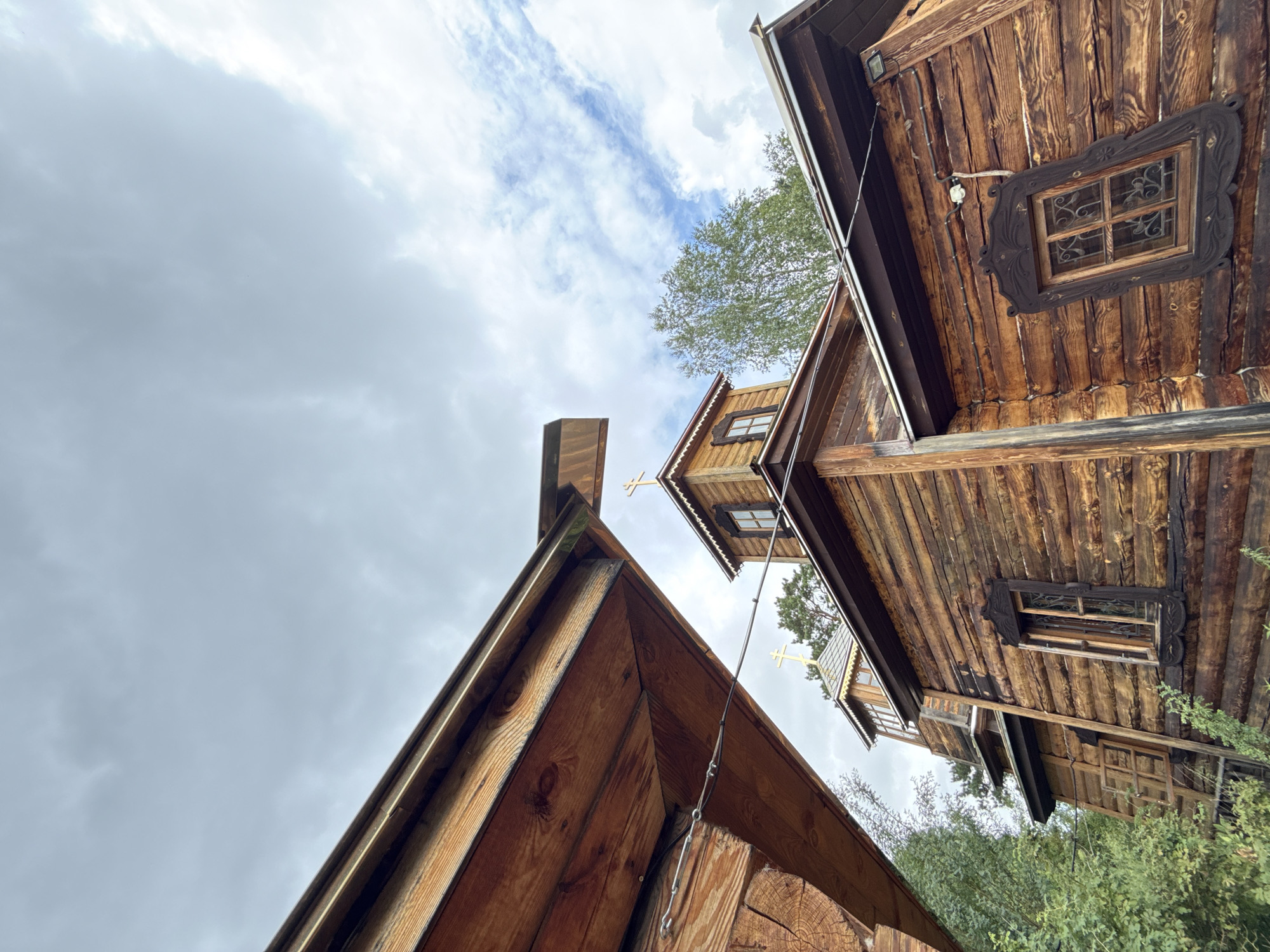 The image shows an exterior view of wooden buildings with Russian Orthodox crosses on their roofs. The buildings are constructed from logs, with visible wood grain and some signs of weathering. One of the structures features a prominent window with an ornate wooden frame. The scene is set against a cloudy sky and surrounded by greenery, suggesting an outdoor location. Based on the architectural style and religious iconography, this is likely a church or monastery in a rural or semi-rural setting. Without specific visual cues to pinpoint the exact location within Chemal, Russia, further details regarding the city cannot be provided. No people or discernible actions are present in the image.