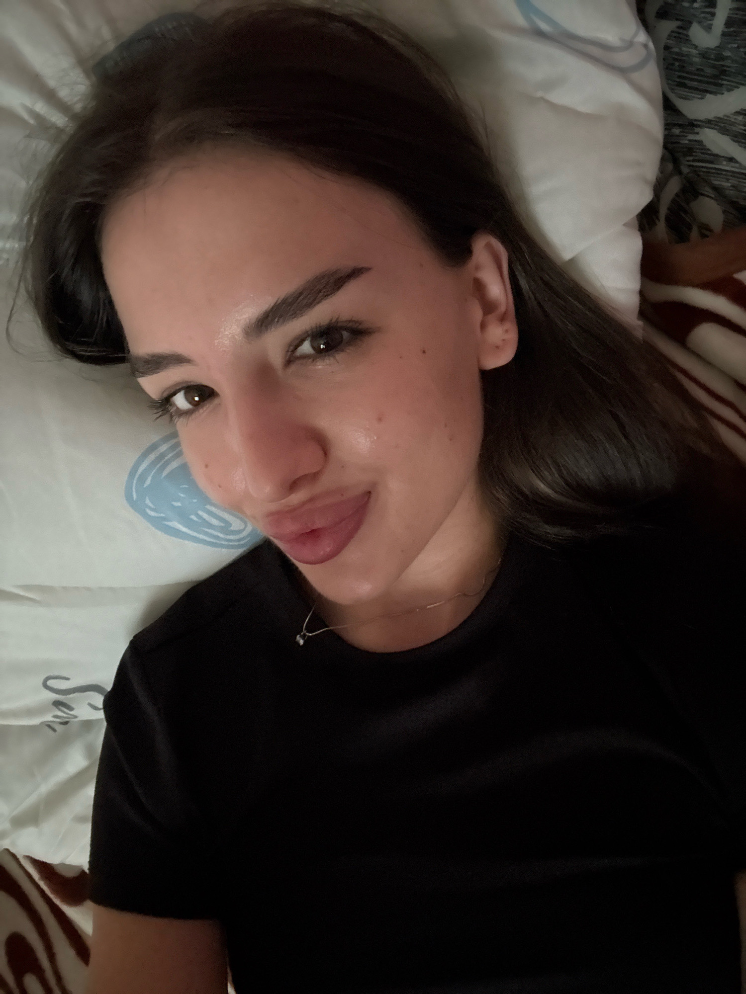 A young woman with long, dark hair and brown eyes is lying on her back on a bed. She is wearing a black t-shirt and a delicate necklace. Her face is turned towards the camera, and she is puckering her lips. The background consists of a white pillow with a blue pattern and a blanket with a brown and white striped pattern. The scene appears to be indoors, likely in a bedroom. There is no indication of the specific location within Basarabeasca, Moldova.