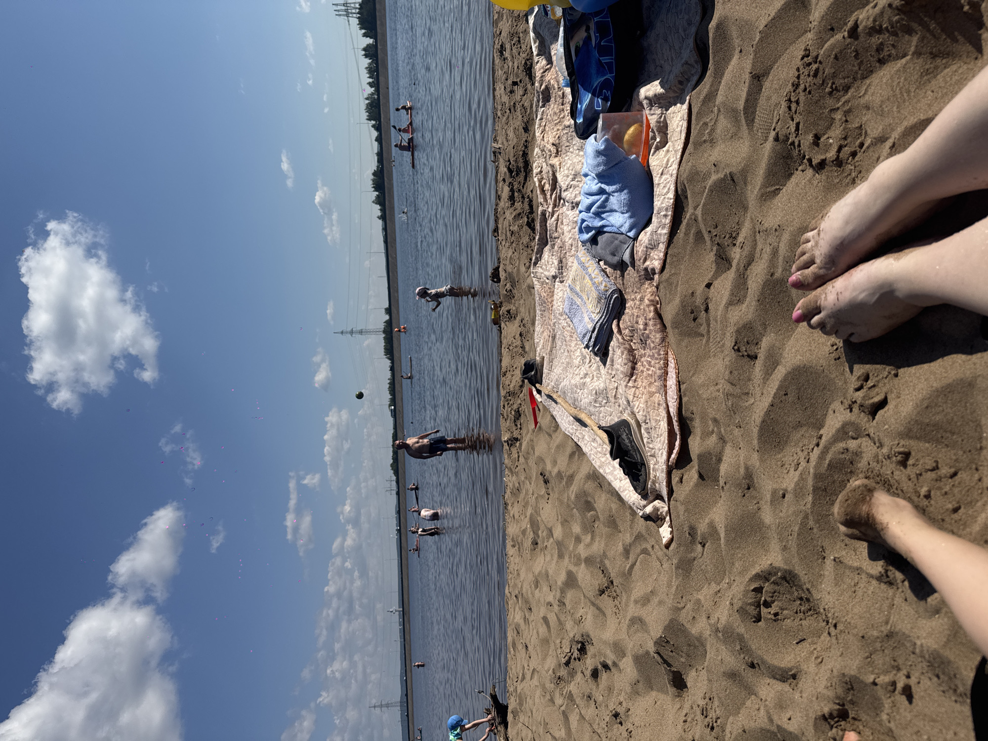 The image depicts a sunny day at a beach in Noviy, Russia. In the foreground, the sandy beach is occupied by a towel with folded towels, a pair of black shoes, and a plastic container with an orange inside. Two pairs of feet are visible in the bottom of the frame, one with painted toenails. In the midground, several people are enjoying the water. Some are wading, while others are on paddleboards. In the background, a body of water stretches to the horizon, with a line of trees and power lines visible on the far shore. The sky is blue with scattered white clouds.
