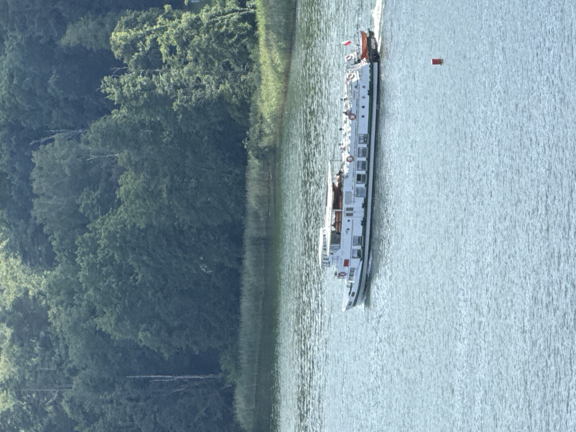The image shows a white passenger boat with several decks, sailing on a body of water. The boat has multiple windows and life preservers visible on its side. A small red and white flag is flying at the stern of the boat, along with what appears to be the number "G02/420" printed on the back. The boat is moving, creating ripples in the water. In the distance, a small red buoy is visible. The left side of the image features a dense green forest with tall trees, suggesting a natural, possibly rural, setting. The water is calm with some texture from the boat's movement and possibly a gentle breeze. The lighting suggests it is daytime, with sunlight illuminating the scene. The overall atmosphere is peaceful and scenic, characteristic of a leisurely boat trip on a lake or river. The location context provided, Mikołajki, Poland, indicates this scene is likely taking place in the Masurian Lake District, a popular tourist area known for its waterways and forests.