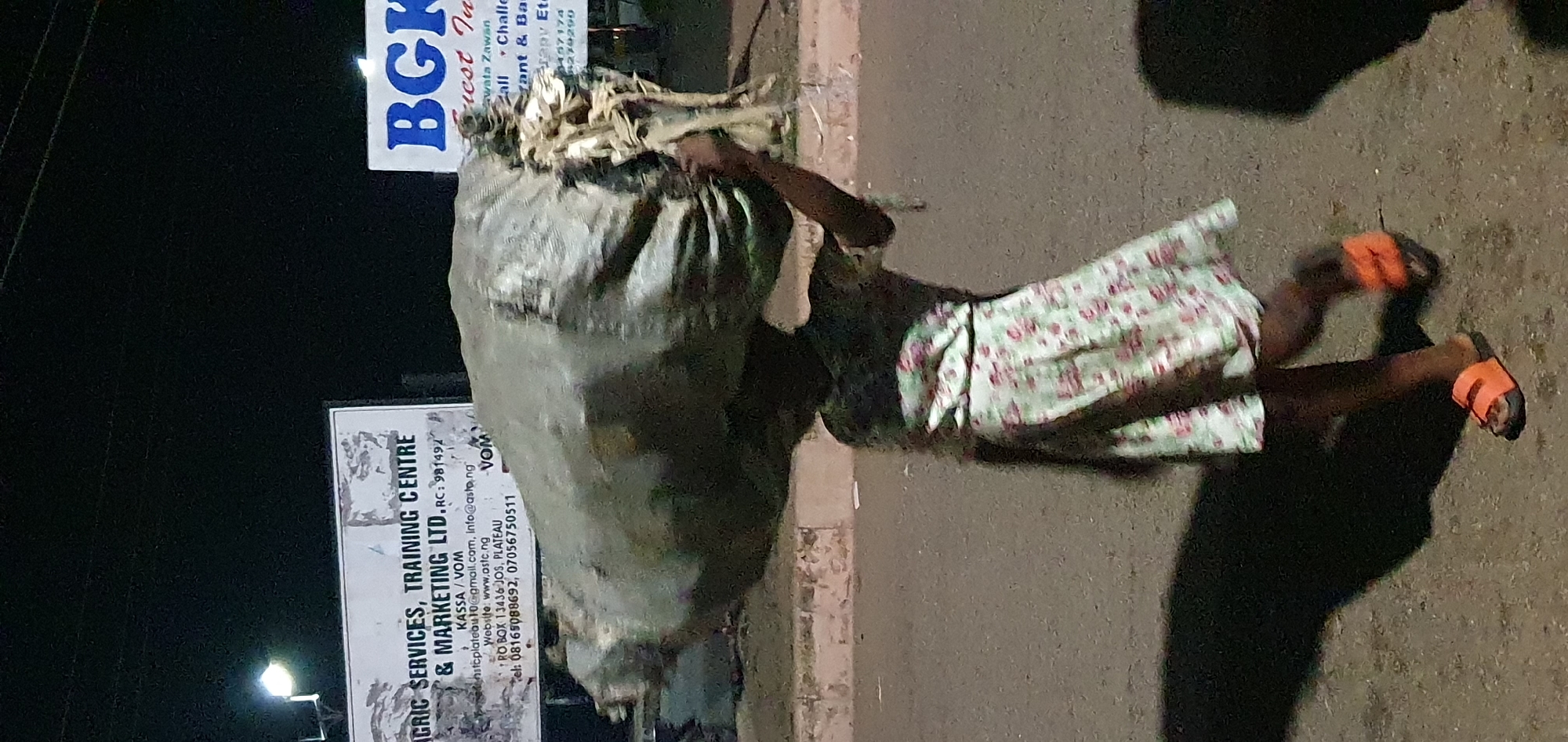 A child is captured in motion, balancing a large, gray, sack-like object on their head. The child is wearing a light-colored dress with a floral pattern and orange sandals. The setting appears to be a street at night in Bukuru, Nigeria, as indicated by the visible signage. One sign reads "AGRIC SERVICES, TRAINING CENTRE & MARKETING LTD." and includes contact information such as an email address and phone number. Another sign, partially visible, displays "BGK". The ground is paved, suggesting a road or sidewalk, and the lighting suggests an outdoor, possibly urban environment. The overall scene conveys a sense of everyday life and labor, with the child engaged in carrying a significant burden.