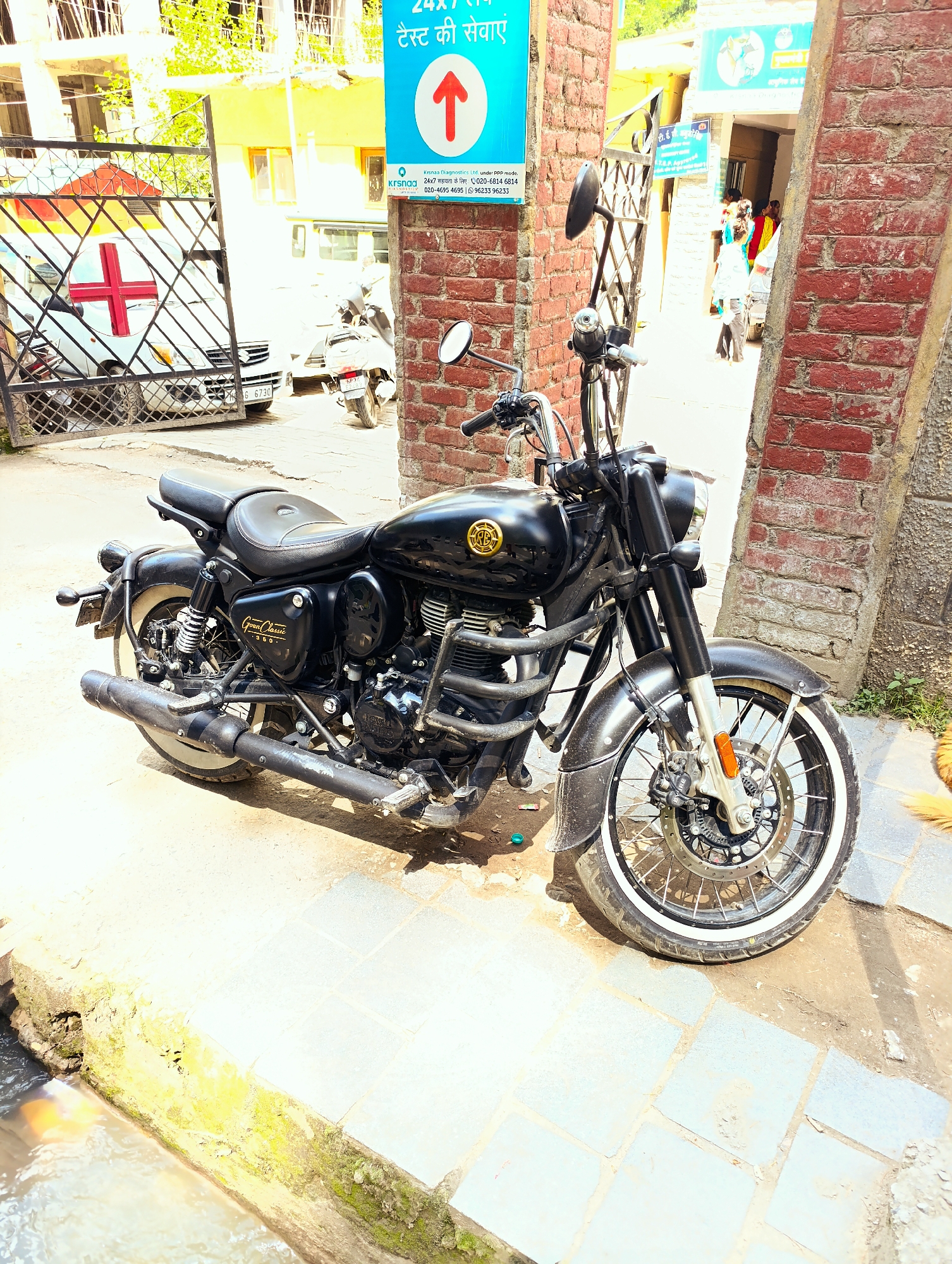 The image shows a black Royal Enfield motorcycle parked on a paved surface next to a brick wall. The motorcycle has whitewall tires and chrome accents. In the background, a white ambulance with a red cross is visible through a metal gate. A blue sign with an arrow and Hindi text is attached to the brick wall. Further in the background, a building with windows and people can be seen. The scene appears to be in a public area, possibly near a hospital or clinic, in Manali, India, given the presence of the ambulance, medical signage, and the style of the architecture. The lighting suggests it is daytime.