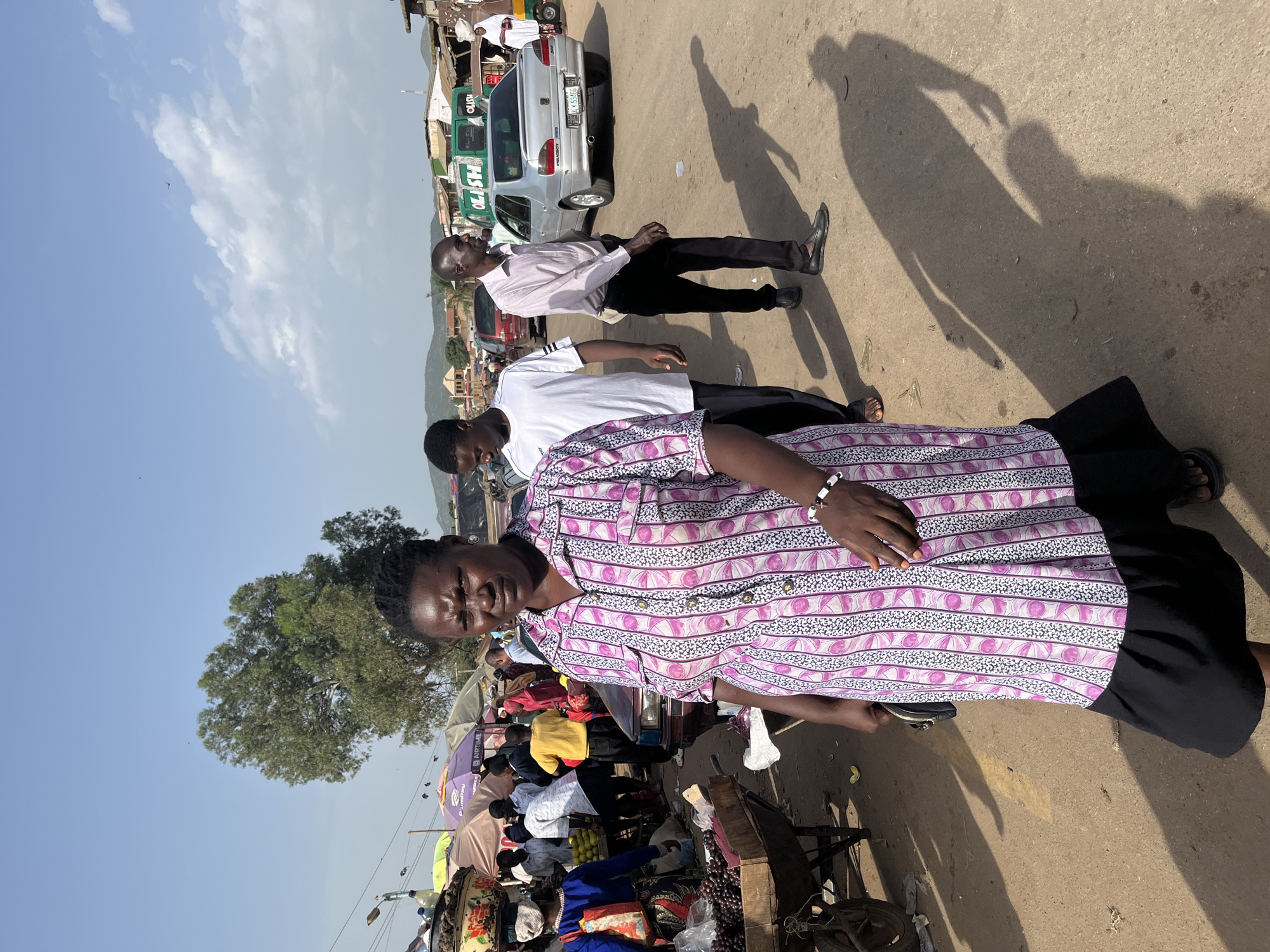 The photograph captures a street scene in Rafin Par, Nigeria, likely a market area. In the foreground, a woman wearing a pink and white patterned dress and a black skirt is walking towards the camera. To her left, a wheelbarrow filled with dark, round fruits, possibly grapes, is visible. Behind the woman, several people are seen walking and interacting. A man in a white t-shirt and another man in a light-colored collared shirt and black trousers are walking away from the camera. A silver Peugeot car is parked on the right side of the road, with a green bus partially visible behind it. The background shows various market stalls with umbrellas and buildings, and distant hills under a clear blue sky with scattered clouds. The ground is a dusty, unpaved road.