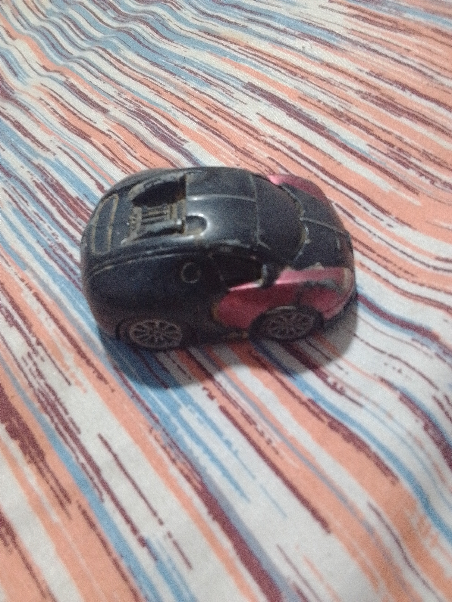 A black toy sports car with pink accents is placed on a striped fabric. The fabric has alternating diagonal stripes of white, light blue, peach, and dark pink. The car is facing towards the left of the frame, with its front slightly elevated. The car appears worn, with some scuffs and scratches visible on its surface. The wheels are silver with a patterned rim. The interior of the car has a black dashboard. The lighting suggests it is daytime, and the setting appears to be indoors or on a covered surface, given the lack of natural shadows and the focus on the car and fabric. There is no visible text in the image.