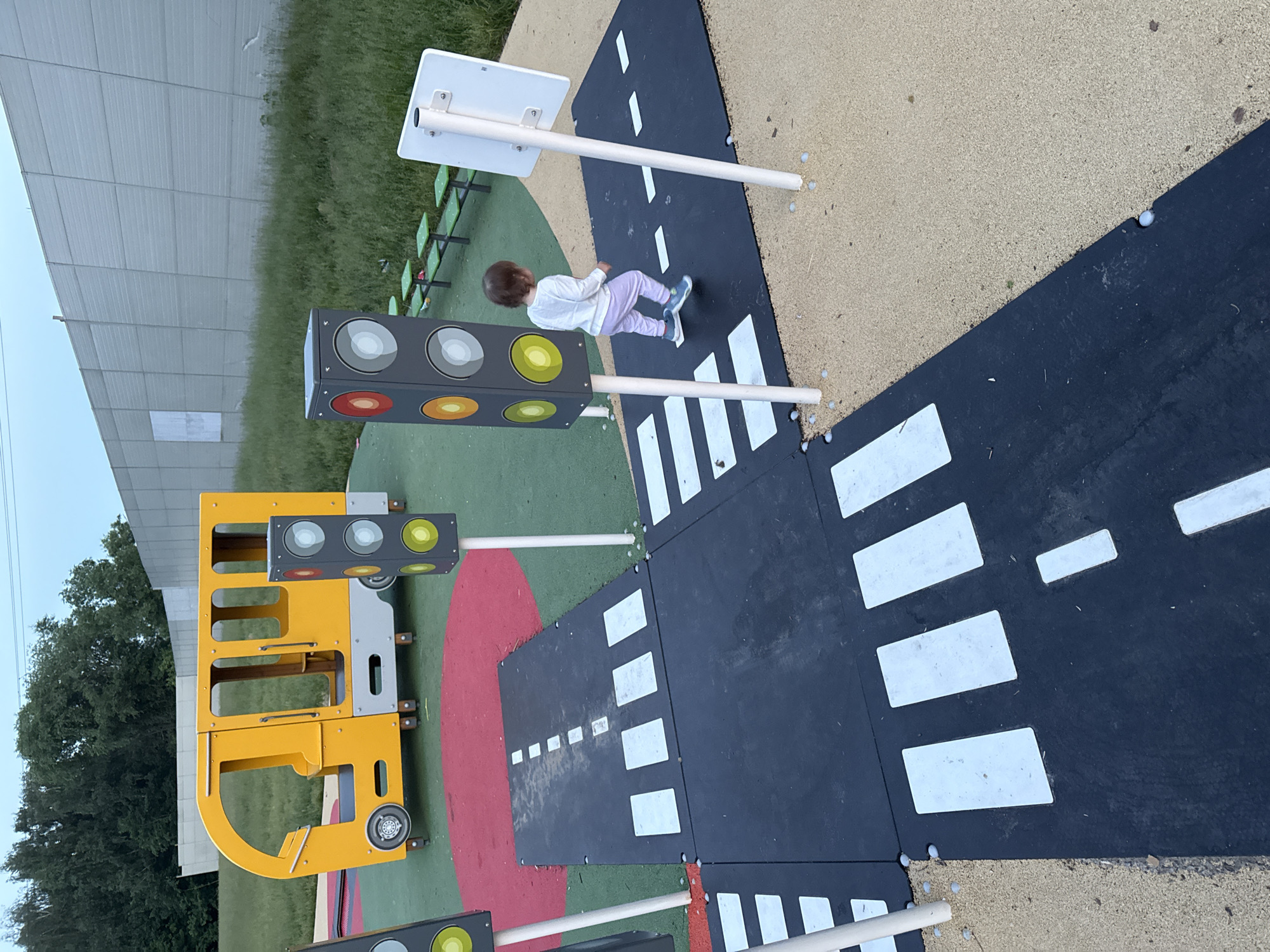 An outdoor play area designed to resemble a city street for children is depicted. A young toddler, seen from behind, walks on a black asphalt-like surface marked with white pedestrian crossings. The toddler is wearing a white long-sleeved shirt and light purple pants. To the left of the toddler stands a grey traffic light structure with colored circles. Further left, a bright yellow and grey play structure resembling a bus is visible. The play area is surrounded by green grass and a tall, light-colored sound barrier wall. In the background, trees and a power line are discernible. The ground is a mixture of green artificial turf and light brown gravel. The image appears to be taken in Moscow, Russia, given the context of other images from the same source.
