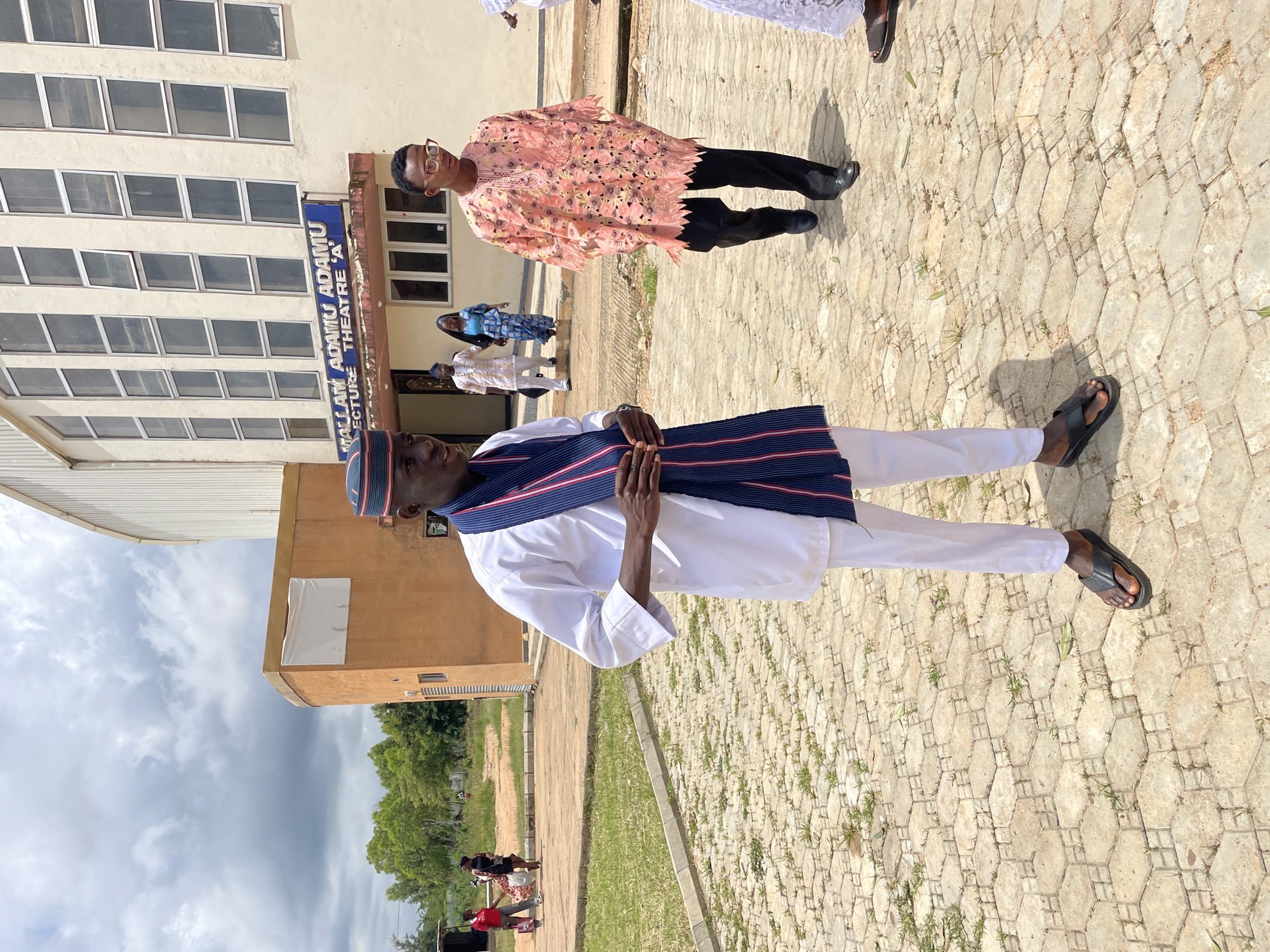 In front of the Mallam Adamu Adamu Lecture Theatre 'A' in Jos, Nigeria, a group of people are gathered outdoors. The primary subject, a young Black man, stands in the foreground, facing right. He is wearing a white kaftan, white trousers, a red, blue, and white striped scarf around his neck, and a matching cap. He is also wearing black flip-flops. Behind him, further into the scene, a man in a pink, intricately patterned robe and black trousers walks towards the foreground. Two other individuals, a man in a white garment and a woman in a blue and white patterned dress, are seen walking away from the viewer towards the entrance of the lecture theatre. In the background, other people are visible, and beyond them, trees and a cloudy sky. The ground is paved with hexagonal interlocking stones.