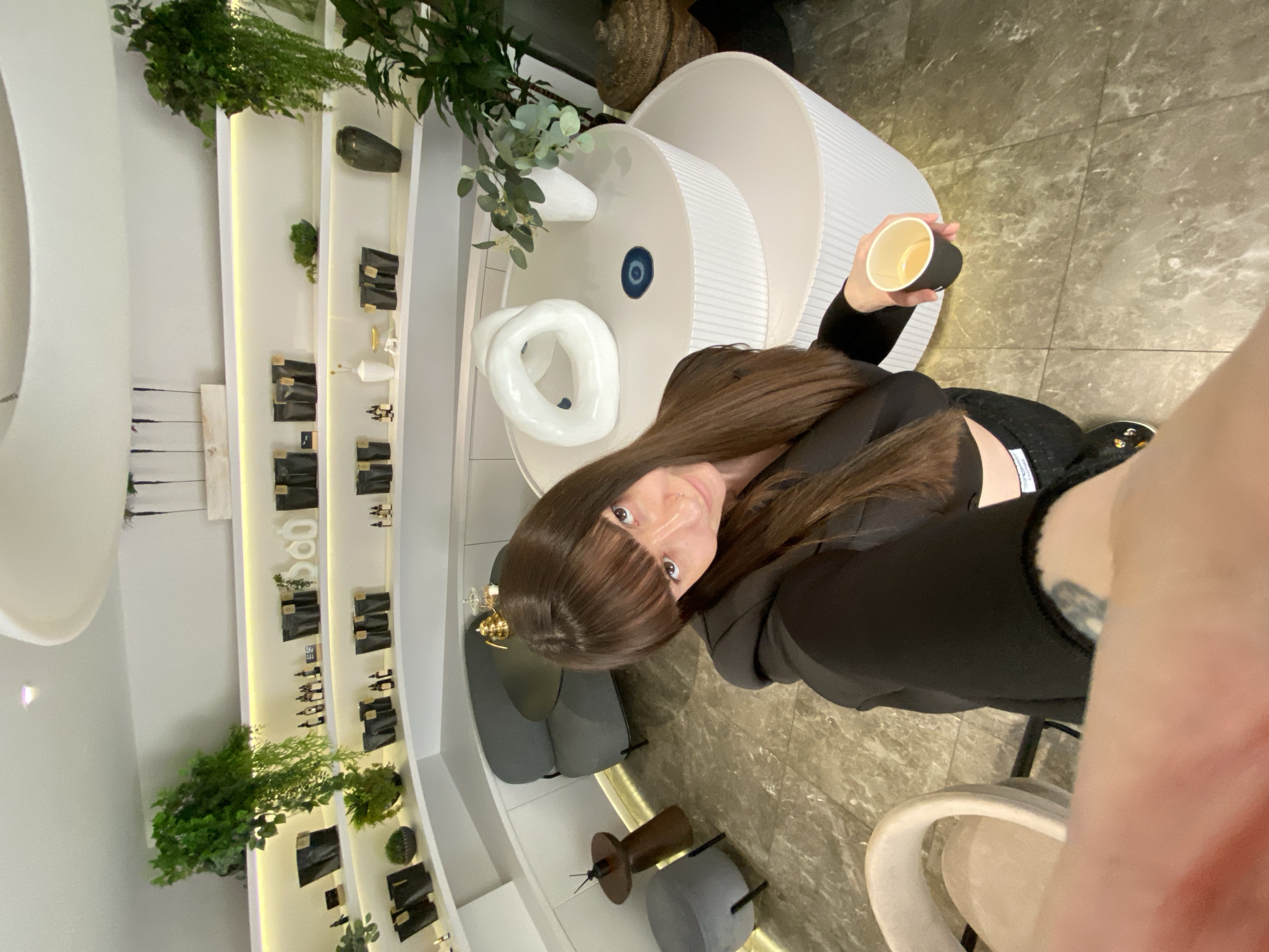A young woman with long, dark brown hair and bangs is captured in a high-angle selfie indoors, likely in Votkinsk, Russia. She looks directly at the camera with a neutral to slightly smiling expression, her eyes visible, along with a nose piercing. She is wearing a black long-sleeved top or hoodie and holds a black paper cup with a light-colored interior, containing a light brown liquid, possibly coffee or tea. Her right arm is visible in the foreground, partially obscuring the view.
The setting appears to be a modern, stylish interior, possibly a cafe, boutique, or showroom. The flooring is a polished tiled surface with a gray and brown marble-like pattern. In the background, there is a large, white, cylindrical, ribbed table or pedestal adorned with a white, abstract knot-like sculpture and a circular blue decorative element. Green plants, including ferns, are strategically placed throughout the space.
Along the wall, white, illuminated display shelves showcase various items. These include numerous black rectangular packages or bags, small bottles (possibly cosmetics or perfumes), and decorative vases of different sizes and colors (one dark, one white), along with some small golden ornaments. Gray upholstered chairs and stools, as well as a small, dark wooden stool with a metal base, are visible as seating options in the background. The overall ambiance is clean, contemporary, and well-lit. No legible text is visible on any of the objects or within the scene.