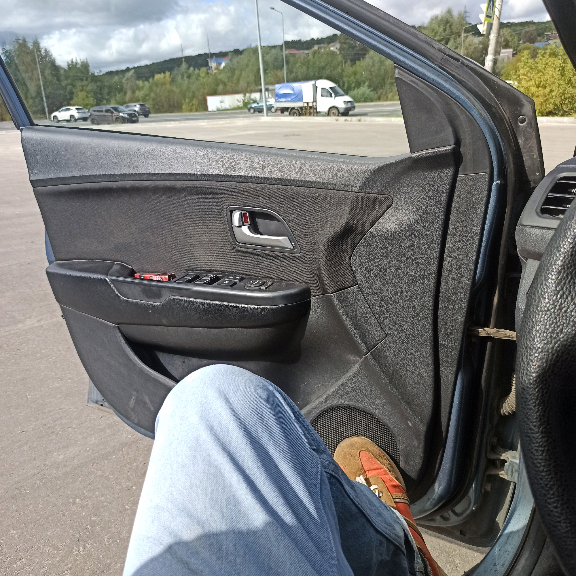 The image is a first-person perspective from inside a car parked on a paved surface, likely a parking lot. The car door is open, revealing the interior door panel, which is black plastic with fabric accents. On the door panel, there is a silver interior door handle and a control panel for windows and mirrors. A small red and silver object, possibly a pack of cigarettes or a lighter, rests on the armrest. The viewer's legs, clad in light blue jeans and a brown and orange sneaker, are visible in the foreground, suggesting they are sitting in the driver's seat.
Outside the car, several vehicles are visible on a road in the background, including a white truck with a blue tarp covering its cargo. The landscape beyond the road features trees and a hill, with a partly cloudy sky overhead. The overall impression is a casual stop in a semi-urban or suburban area. Without further context, it is impossible to definitively place this scene in Nizhny Novgorod, Russia.
