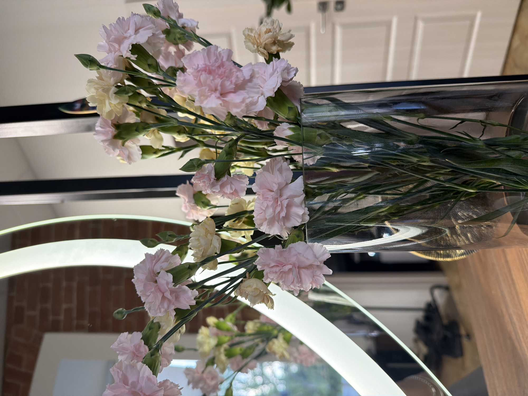 The image shows a clear glass vase filled with a bouquet of pink and pale yellow carnations. The vase is placed on a wooden surface, likely a table or shelf. Behind the vase, a large, vertically oriented mirror reflects the flowers and the surrounding environment. The reflection reveals a brick wall, a white door with paneling, and a circular illuminated light fixture integrated into the mirror's frame. The lighting suggests an indoor setting, possibly a home or a commercial space. There are no people or animals visible in the image. The location of Buzuluk, Russia, is not determinable from the visual information provided in the image.