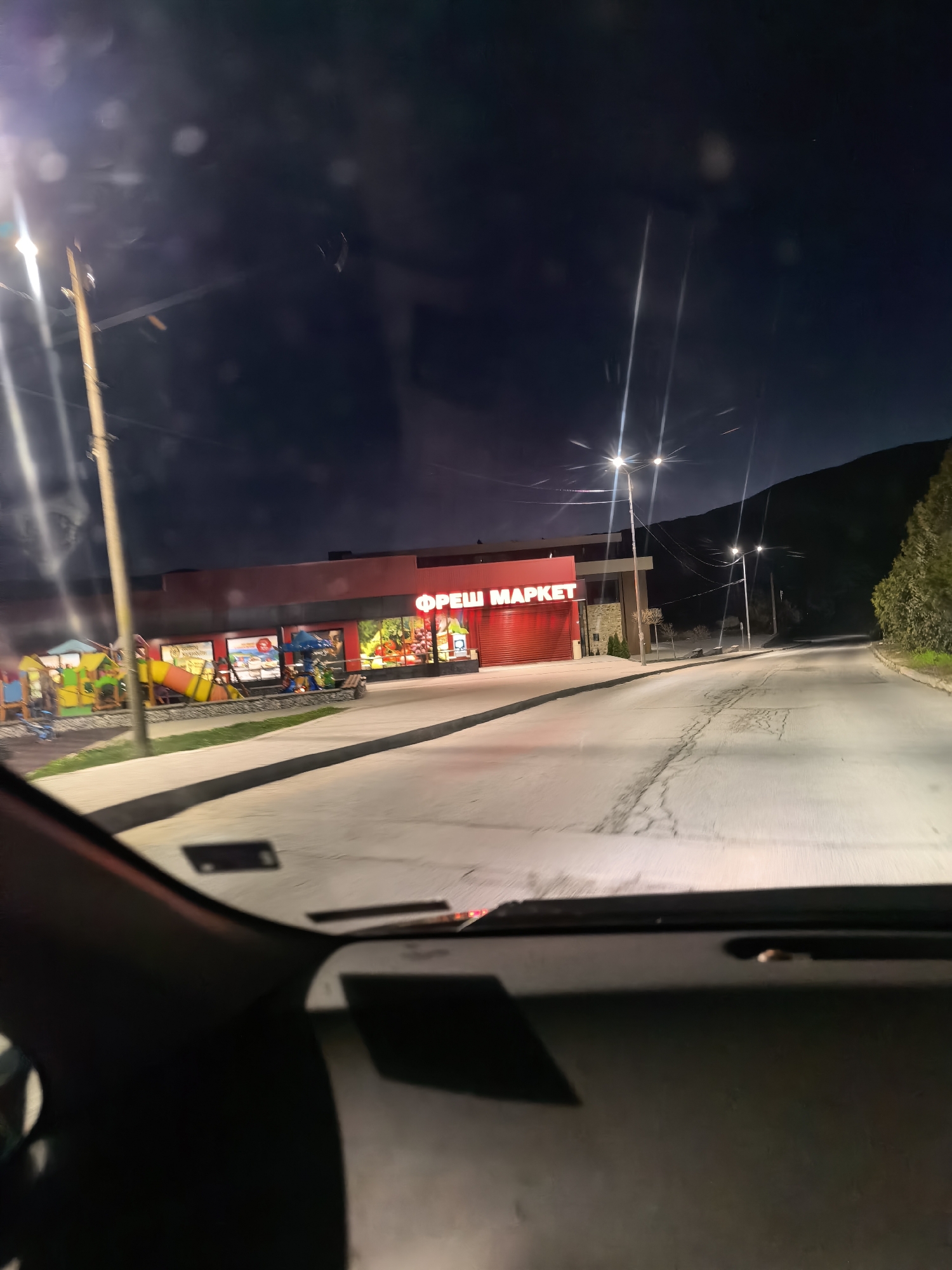 The image shows a view from inside a car at night, looking out at a "ФРЕШ МАРКЕТ" (FRESH MARKET) in the city of Unknown, Unknown. The market is brightly lit, with a red sign above it. There is a playground with colorful equipment to the left of the market. Streetlights line the road leading away from the market, and a dark hill or mountain can be seen in the background. The car's dashboard and windshield are visible in the foreground. The road appears to be paved.