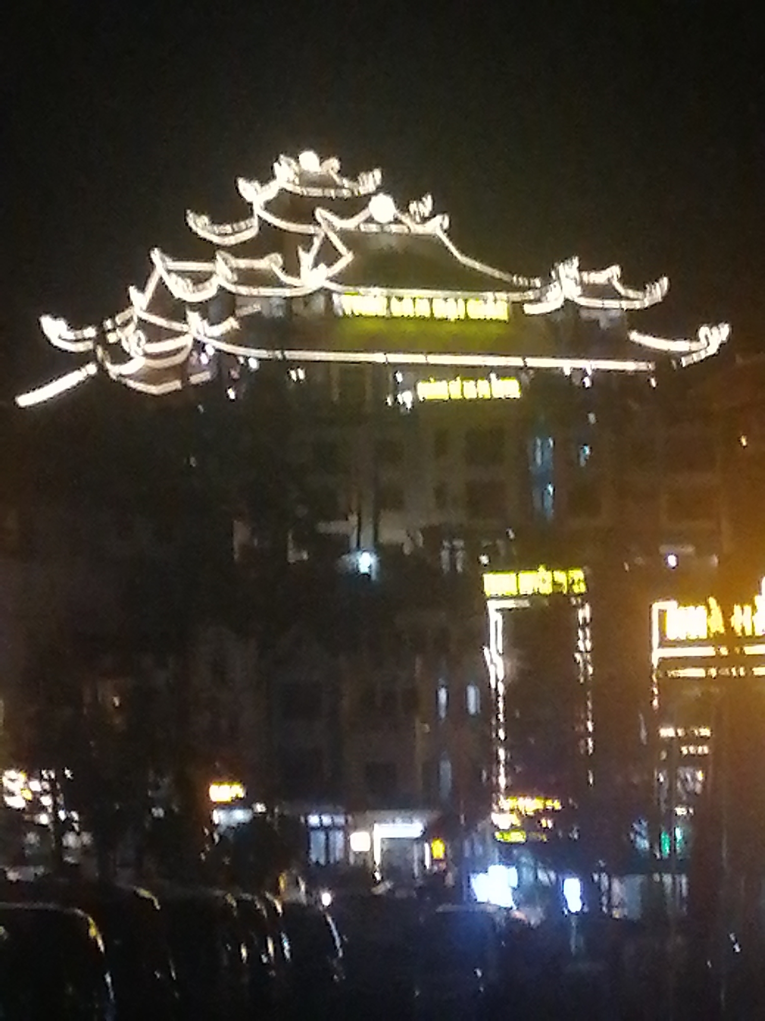 At night, a building with a traditional Vietnamese roofline is illuminated by string lights. The building appears to be a hotel, with multiple floors and lighted signage. Below, a street scene includes parked cars and other businesses, also lit up at night. The location is most likely in Sa Pa Ward, Vietnam, given the architectural style. No people are clearly visible.