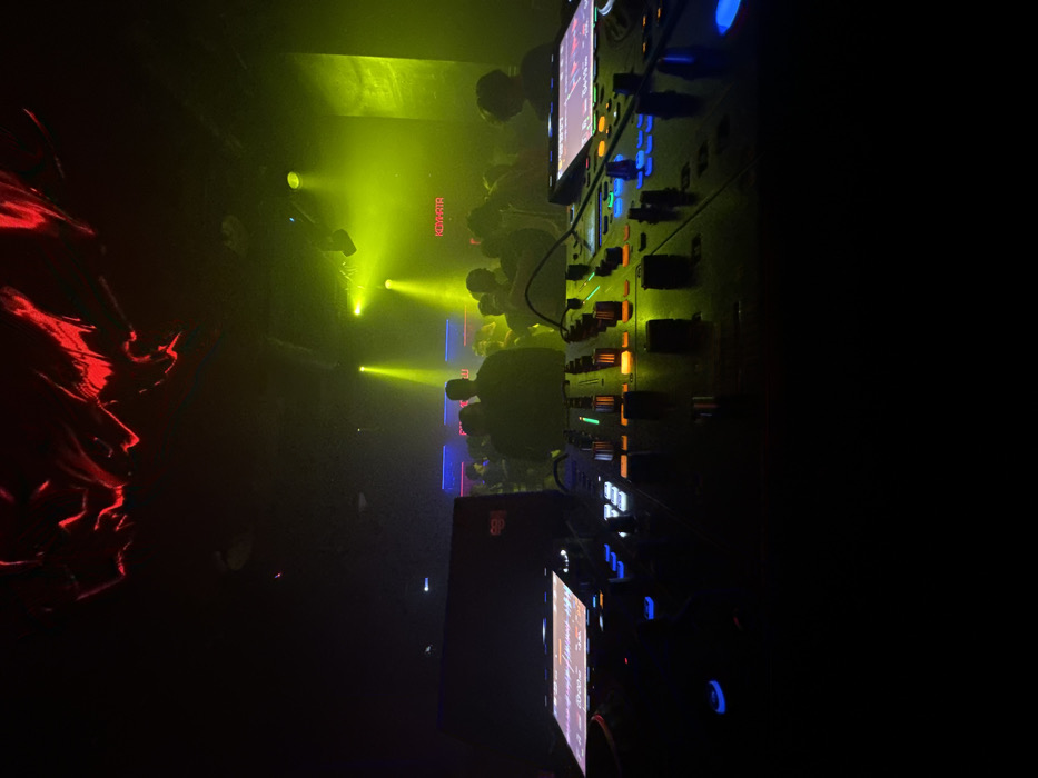 A dark, blurry scene inside what appears to be a nightclub. A crowd stands bathed in yellow light, with a sign reading "KOYATA" visible. To the right, a DJ console with various controls and screens glows with multiple colors. Red lighting creates a distinct shape on the left, possibly fabric or a decorative element. The overall impression is a lively, crowded, and energetic atmosphere focused on music and entertainment.