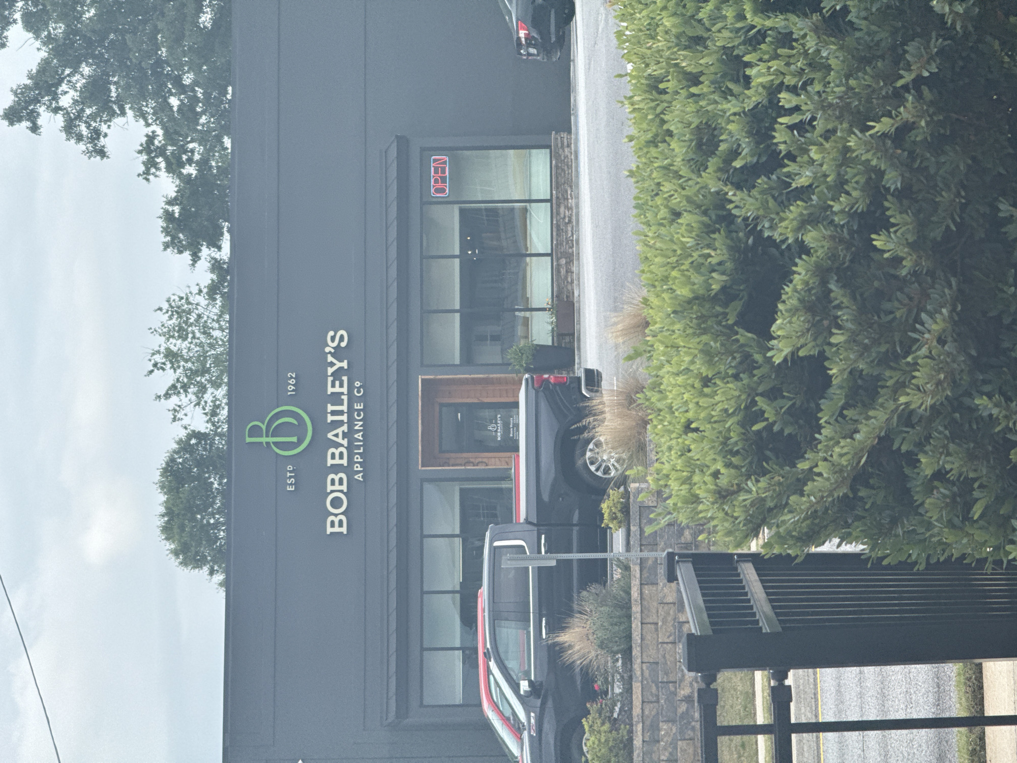 The image depicts the storefront of "Bob Bailey's Appliance Co." in Stone Mountain, USA. The building is a dark gray, one-story structure with a prominent logo and signage. An "OPEN" sign is visible in the window. Two dark-colored pickup trucks are parked in front of the store. Landscaping elements including bushes and decorative grasses are also visible. The weather appears overcast.