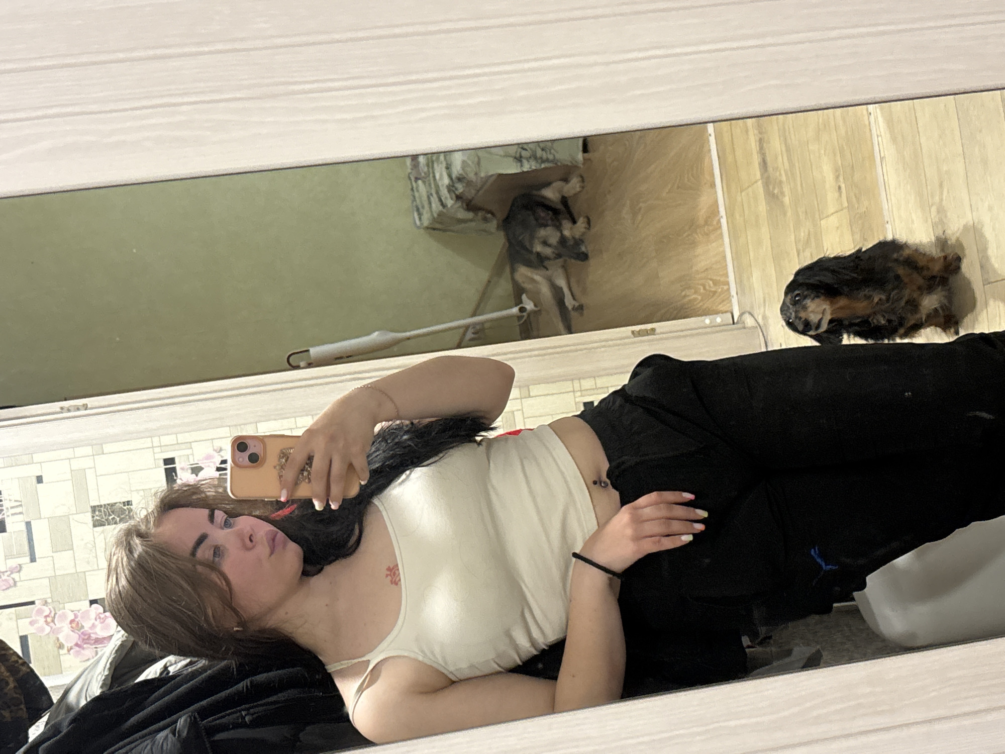 A woman is lying on a bed in what appears to be a room in Izhevsk, Russia, taking a selfie in a mirror. She is wearing a white tank top and black pants. Two dogs are visible in the reflection. The room has a tiled wall with a floral pattern, and there is laminate flooring. One dog is sitting near a pet bed or elevated platform, and the other is lying on the floor.