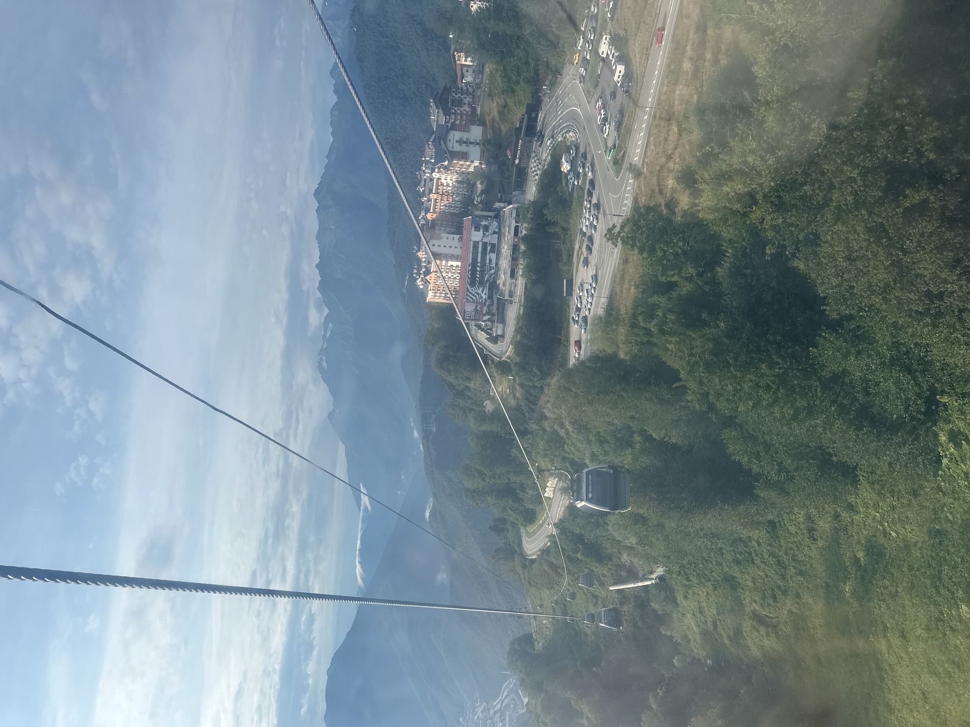This image shows a view from a cable car in the city of Estosadok, Russia, in the Caucasus Mountains. The cable car is moving upwards, revealing a panoramic view of the surrounding landscape. In the foreground, lush green trees cover the steep slopes of the mountains. A winding road with several cars can be seen snaking its way up the mountainside, leading to a complex of buildings that appear to be hotels or resorts. Further in the distance, more cable cars are visible, suspended on thick steel cables that stretch across the frame. The sky is a pale blue, dotted with fluffy white clouds, and the distant mountains are hazy and blue in the soft light. The scene suggests a popular tourist destination, with its well-developed infrastructure for enjoying the natural beauty of the area.