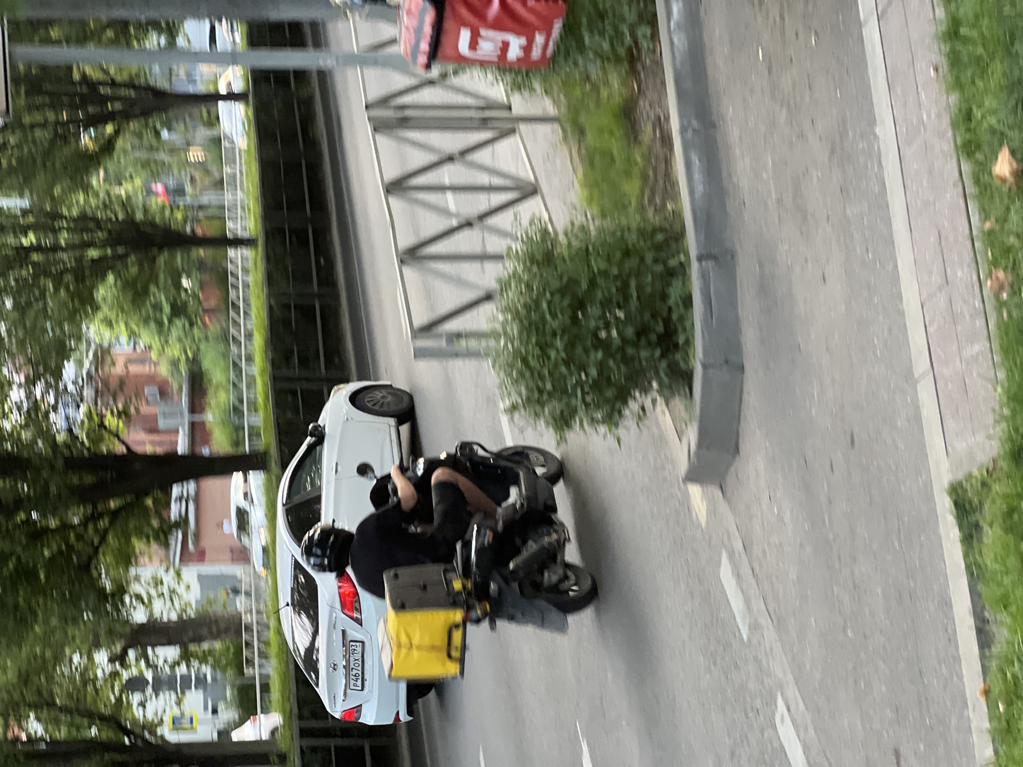 A male individual wearing a black helmet and black clothing is riding a black scooter with a yellow delivery box on the back. The scooter is traveling on a paved road, moving away from the camera. To the left of the scooter, a white car with a license plate that reads "P467OX 193" is also moving away from the camera. The background consists of trees, buildings, and a metal fence. The scene appears to be in an urban environment, possibly in Krasnodar, Russia, given the context of the provided information.