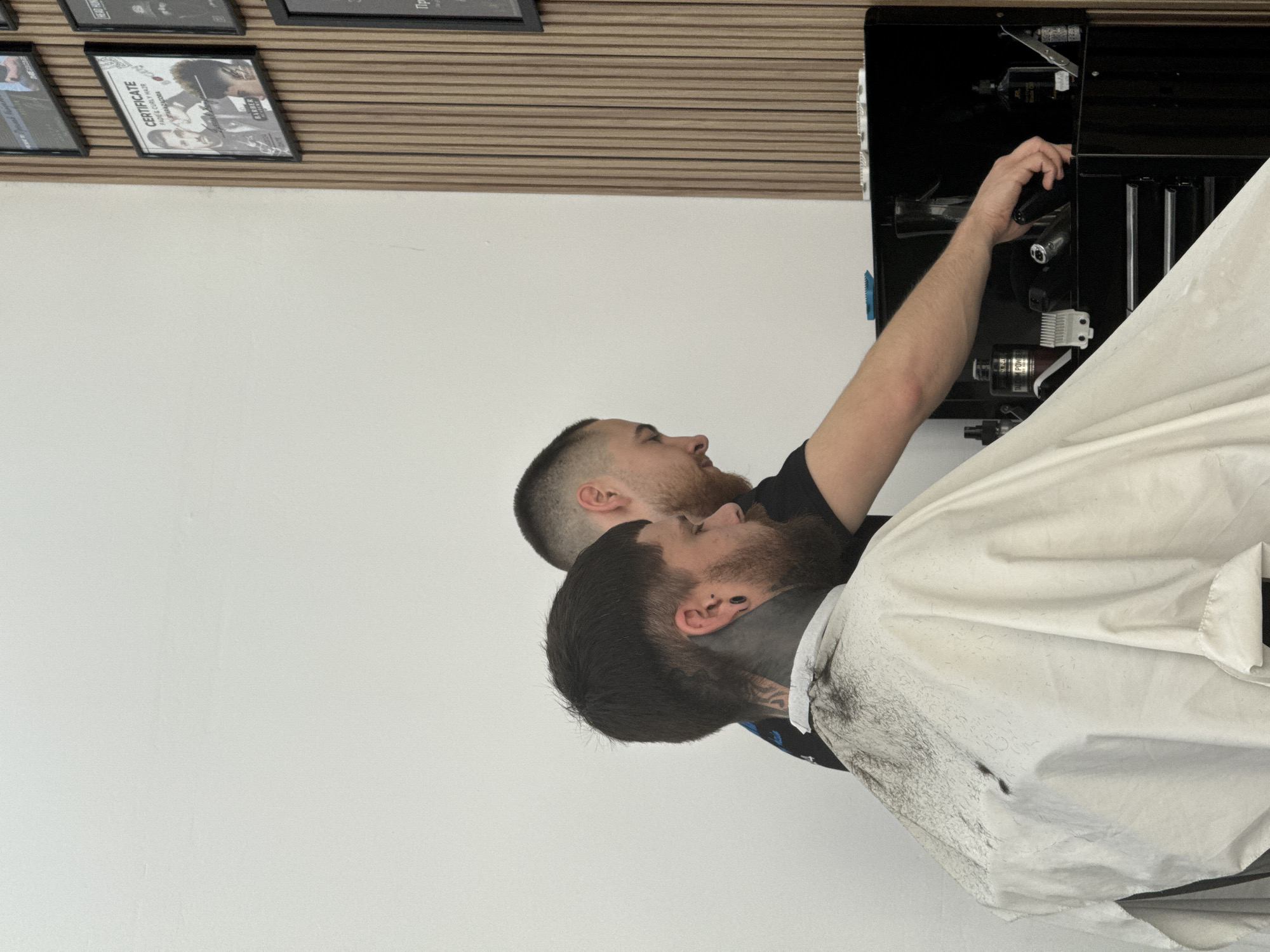 An indoor scene in a barbershop in Sofia, Bulgaria, is captured in a sideways (rotated 90 degrees clockwise) orientation. The image shows a barber giving a haircut to a client.
In the center, two men are visible in profile. The barber, positioned on the right side of the frame (if viewed upright), has short dark hair and a beard, and wears a dark shirt. His right arm is extended upwards, reaching into a black, wall-mounted organizer or cabinet that holds various barber tools such as clippers and trimmers.
The client, seated to the left of the barber, has dark hair, a beard, and visible tattoos on his neck below his ear. He is draped in a white protective cape that is speckled with dark hair clippings, indicating a haircut is either underway or has recently concluded. Both individuals appear focused on the activity.
The background features a two-toned wall. The upper left portion consists of light brown, horizontally oriented wooden slats. Several framed pictures or certificates are hanging on this wooden section. One framed document clearly displays the word "CERTIFICATE" and "ALREX," with other smaller text that is difficult to decipher. The lower left portion of the wall is plain white. The overall lighting suggests an indoor setting.