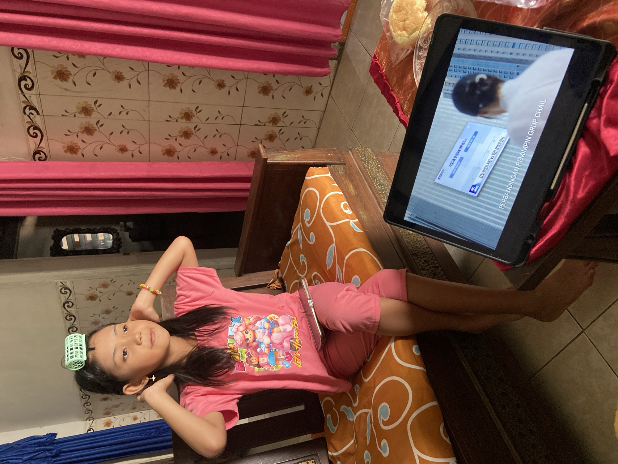 In a residential setting in Baron, Indonesia, a young girl with a hair curler on her head sits on a wooden bench with an orange and white patterned cushion. She is wearing a pink t-shirt with cartoon characters and pink shorts. A mobile phone rests horizontally across her lap. Her hands are raised to her ears, and she has a beaded bracelet on her right wrist. In the foreground, a tablet computer displays a screen with Korean text and an image. A slice of bread is visible on a plate to the right of the tablet. The background features pink curtains and floral patterned wallpaper.