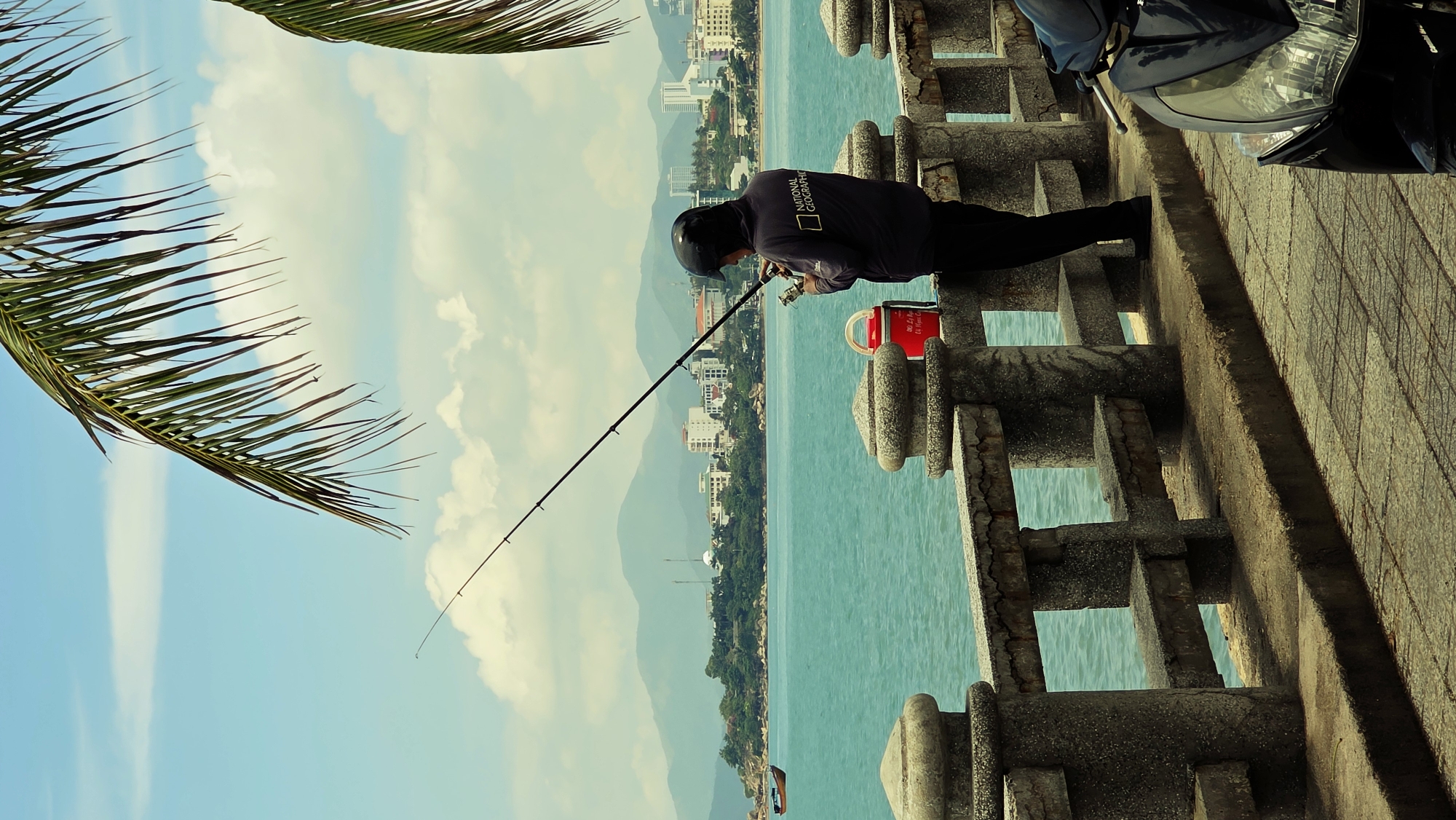 A man is fishing off a concrete pier or walkway with a decorative railing. He is wearing a dark long-sleeved shirt with "NATIONAL GEOGRAPHIC" written on the back, dark pants, and a helmet. The man is holding a fishing rod and appears to be focused on the water. A red container is next to him on the pier. A black motorcycle is partially visible on the right side of the frame. In the background, there is a coastal cityscape with buildings, trees, and hills, likely Nha Trang. The water is a light blue-green color. A palm frond is partially visible in the upper left corner of the image. The sky is blue with scattered clouds.