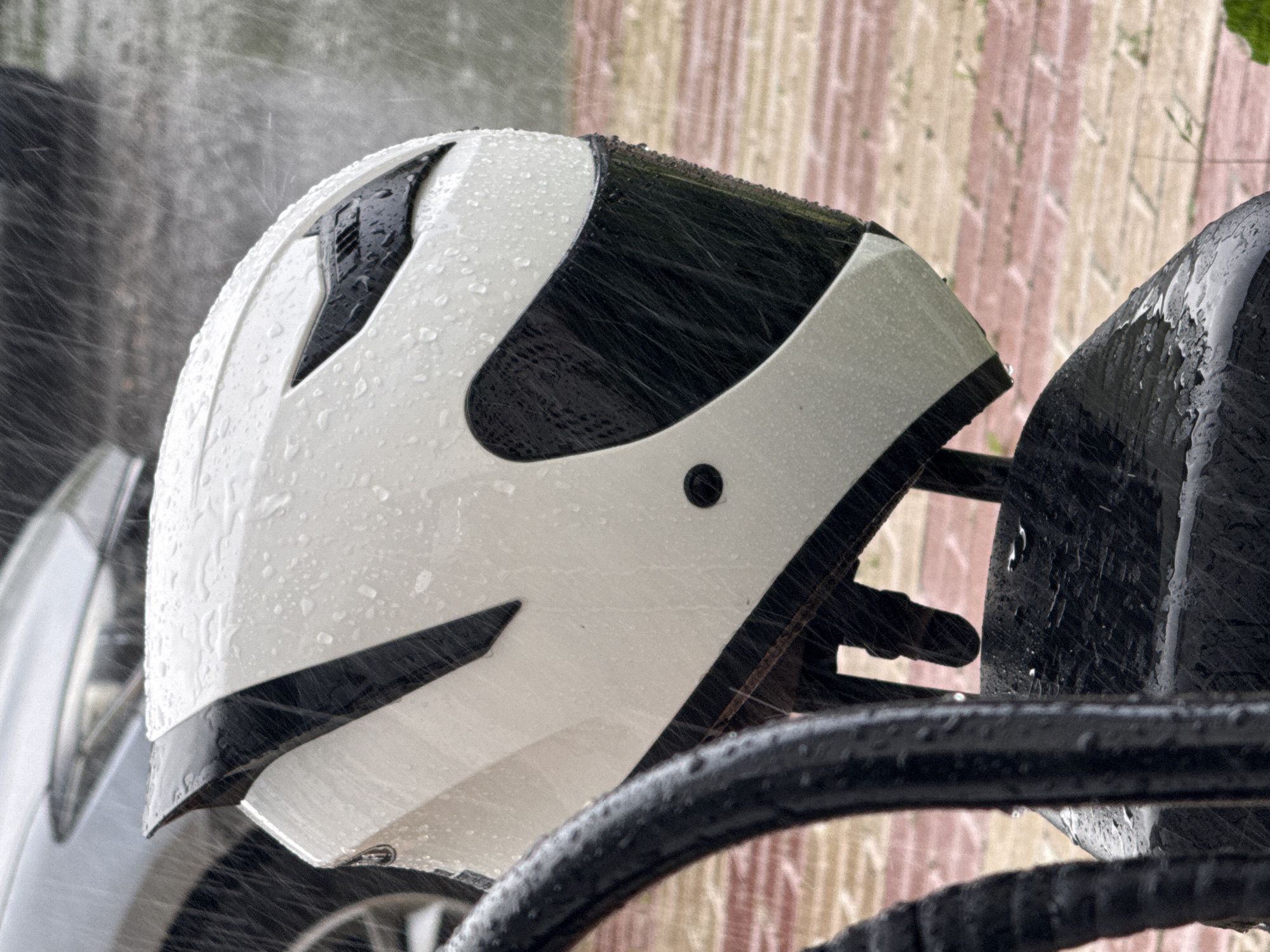 A white and black motorcycle helmet is depicted outdoors in the rain. The helmet is wet with raindrops and is resting on a dark, textured surface, possibly a motorcycle seat. In the background, a blurred vehicle and a reddish-brown tiled ground are visible, with rain streaks suggesting inclement weather. The scene appears to be in an urban environment, but there is no specific indication of its location in Moscow, Russia.