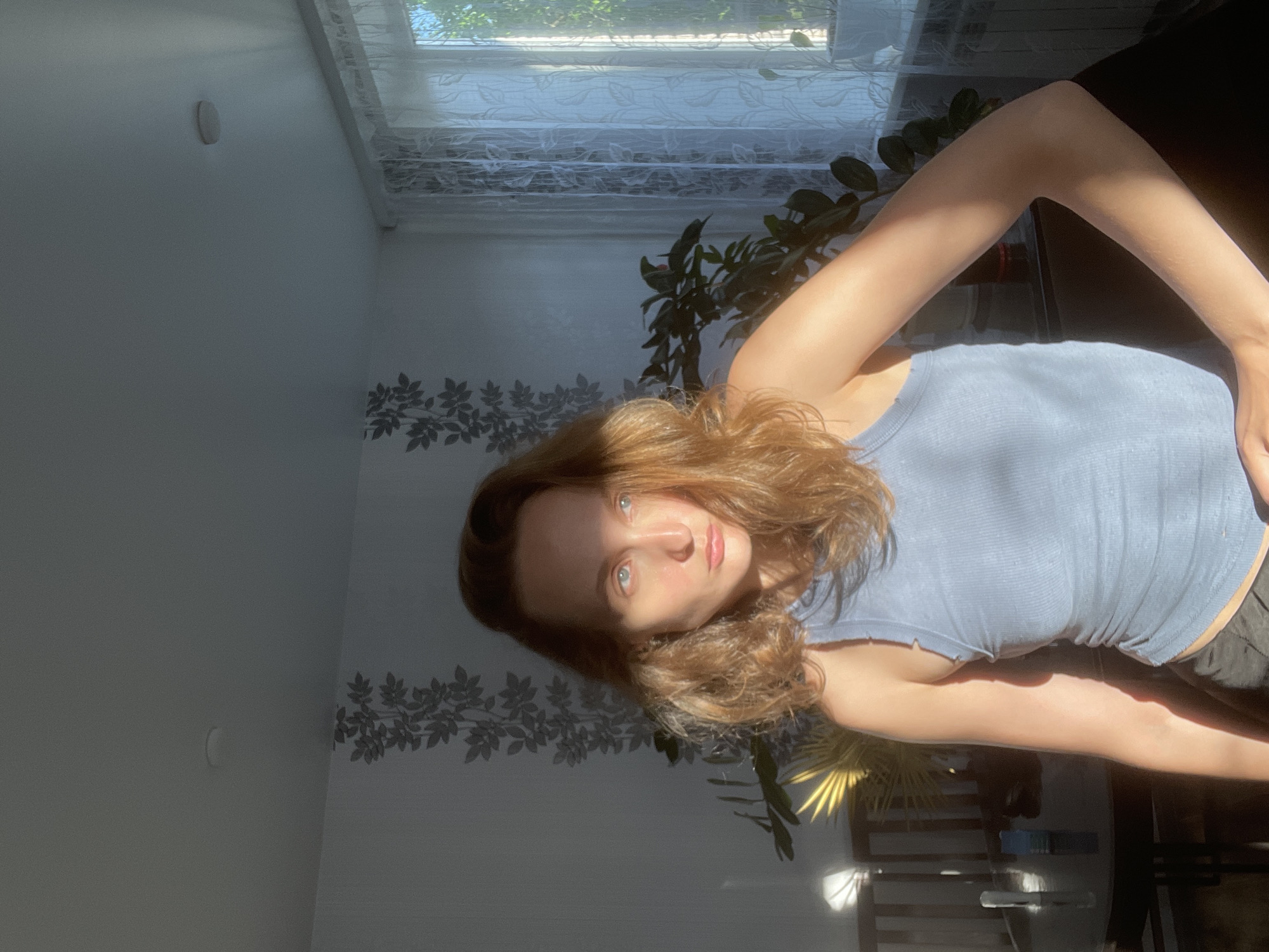 The image captures a young woman in an indoor setting, likely a portrait or selfie, bathed in strong natural light. The woman, with shoulder-length, wavy, reddish-brown hair and blue eyes, looks directly at the camera with a calm, neutral expression. She is wearing a light blue ribbed tank top, and her arms are partially visible, posed in a bent position.
Direct sunlight streams in from a window to the right, creating dramatic highlights on the woman's face and arms, and casting distinct shadows across the room. The background features a light-colored wall decorated with a repeating pattern of dark leaf or vine motifs along its lower half. The window, partially covered by a sheer white curtain with a subtle pattern, reveals green outdoor foliage and a bright sky, suggesting it's daytime.
Several indoor plants are visible, adding to the domestic atmosphere; one with large, dark green leaves is positioned behind the woman, and another with lighter, possibly yellowish leaves, is illuminated by the sun in the lower right of the frame. A small, light-colored oval object is affixed to the upper left section of the wall. The strong, angled sunlight indicates it is either morning or late afternoon. The scene depicts a comfortable home environment, likely a living room or bedroom, in Sharkan, Russia. There is no visible text in the image.