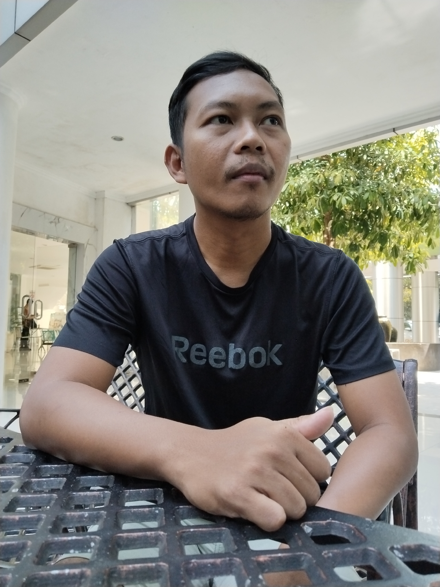 A man with short dark hair and a mustache is sitting at a black metal table outdoors. He is wearing a black t-shirt with the word "Reebok" in gray lettering. His arms are resting on the table, and his hands are clasped. The background shows a building with large glass doors and columns, and a tree with green leaves. The lighting suggests it is daytime. The man is looking up and to the right.
