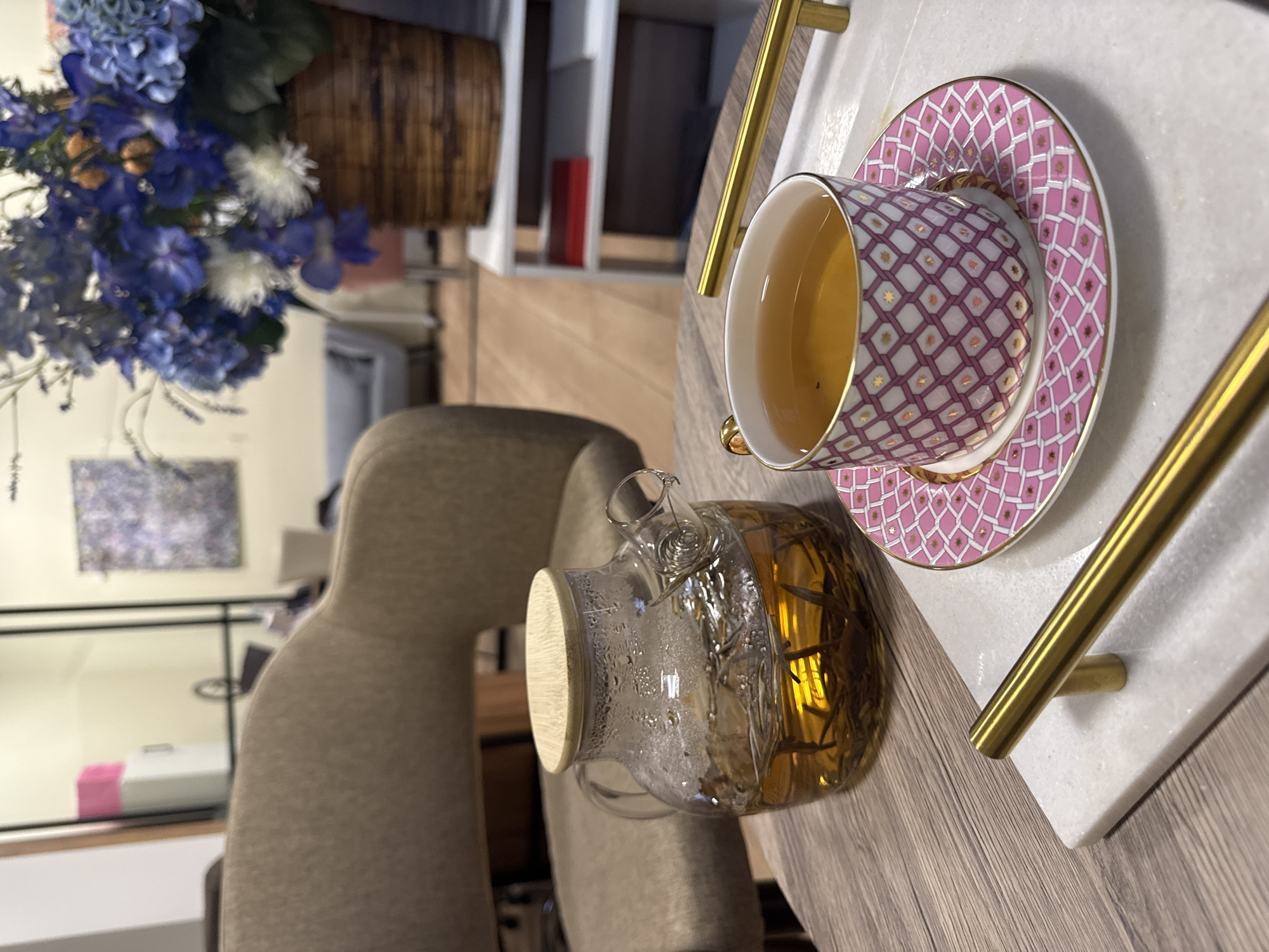 A teacup and saucer with a pink and white geometric pattern, filled with tea, are placed on a white marble tray with gold handles on a wooden table. A glass teapot with a wooden lid sits next to the cup. A bouquet of blue and white flowers is positioned to the left. A beige chair is visible in the background. The background also shows a bookcase, a painting, and a white cabinet. The location is not identified in the city of Moscow, Russia.