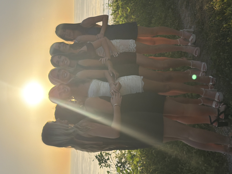 A group of at least seven young women stand in a line, facing forward, with the setting sun to their left. They are wearing dresses of varying colors and styles, along with sandals. They all have their arms folded in front of them. The scene is outdoors, with greenery visible behind them and what appears to be a body of water in the distance. The sun's glare obscures some details and creates a strong highlight on the left side of the image.