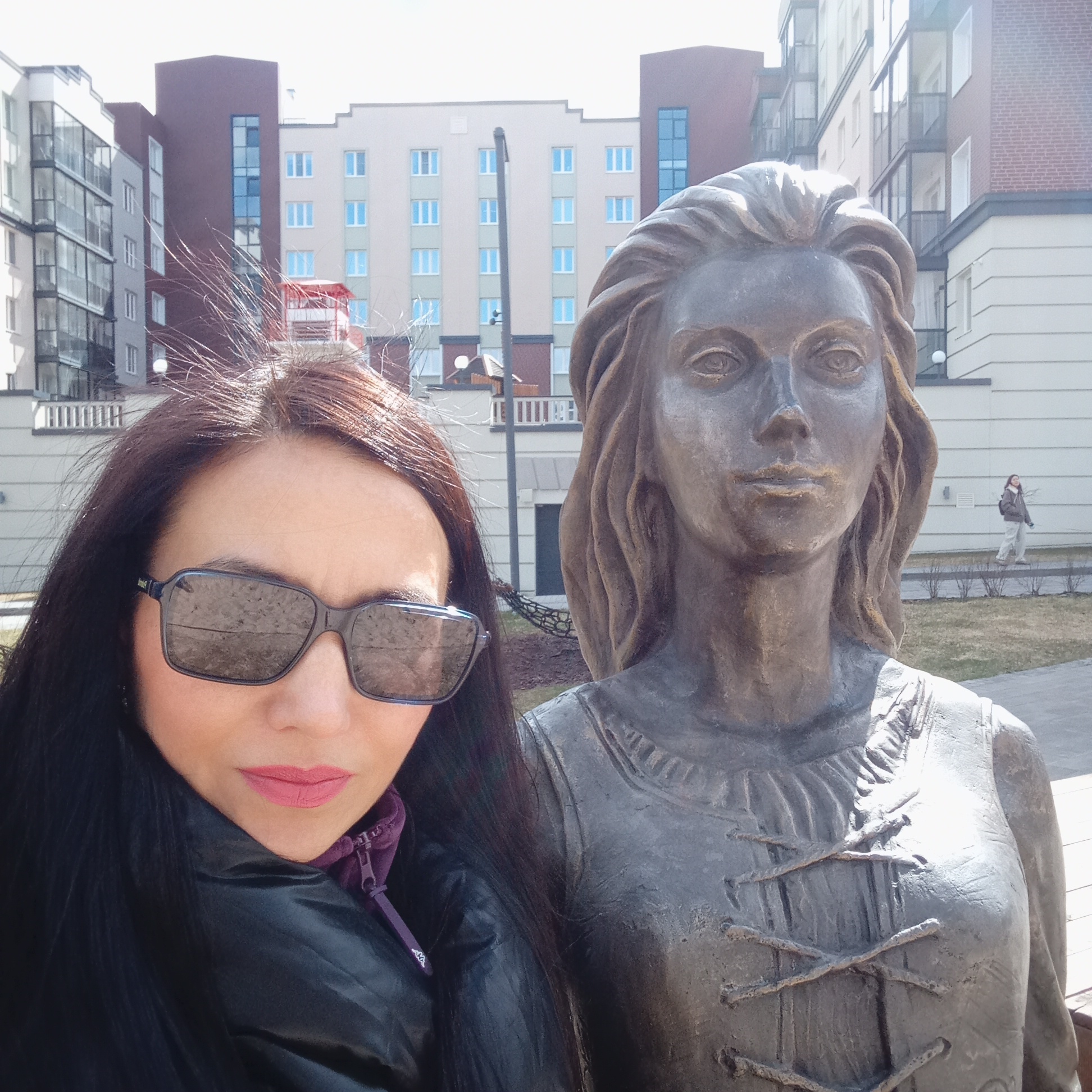 A woman with long dark hair and sunglasses stands next to a bronze statue of a woman. The subject is outside, in an urban area with buildings in the background. The buildings are multi-story and appear to be residential apartments. In the background, a person is walking along a sidewalk. The location appears to be in the city of Pervomaysky, Russia. The woman is wearing a black jacket with a purple zipper and collar, and she has pink lipstick. The bronze statue has detailed features including hair and clothing that is tied together.